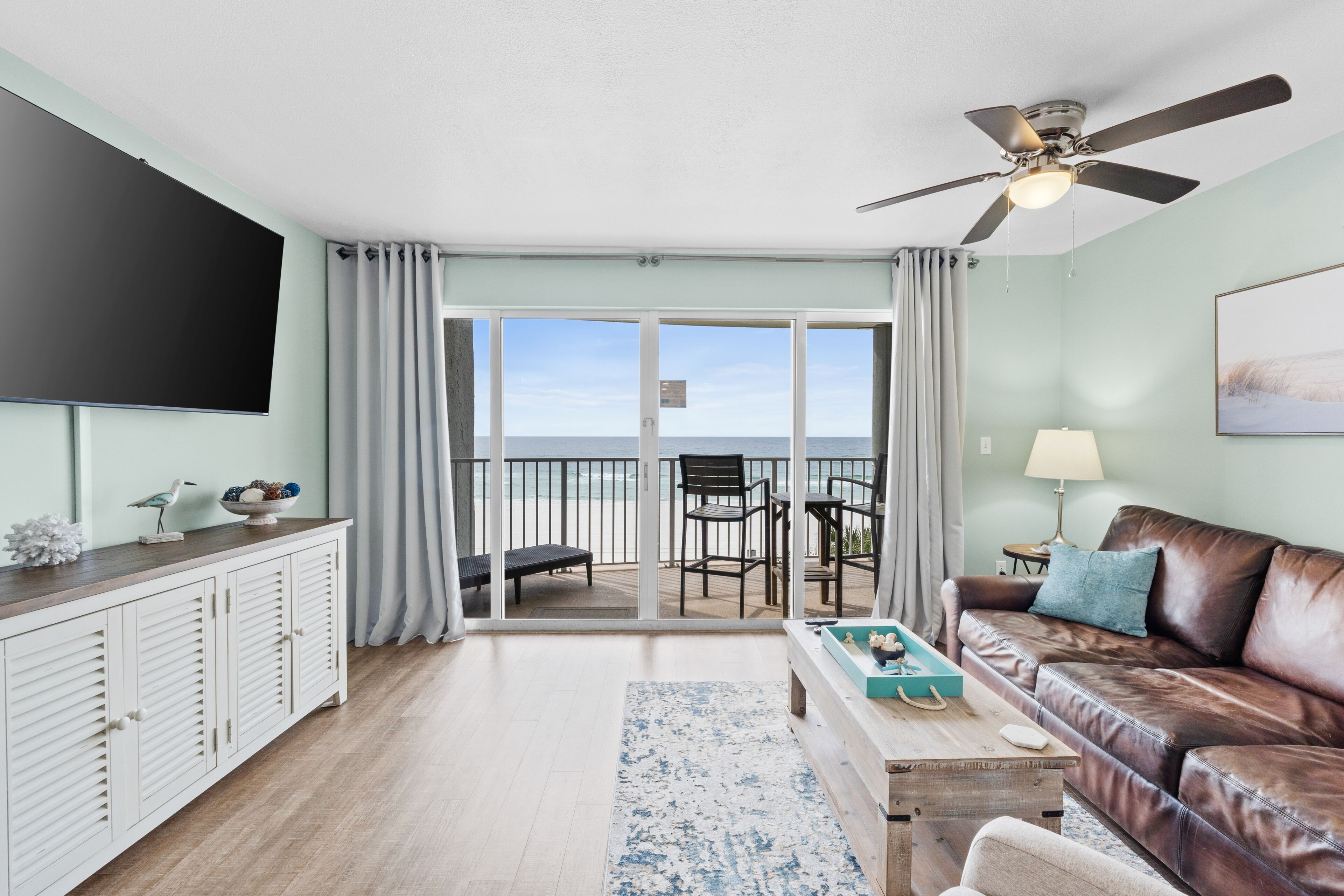 A living room with a view to the ocean, a ceiling fan, a flat-screen TV, a leather sofa, and a wooden coffee table.
