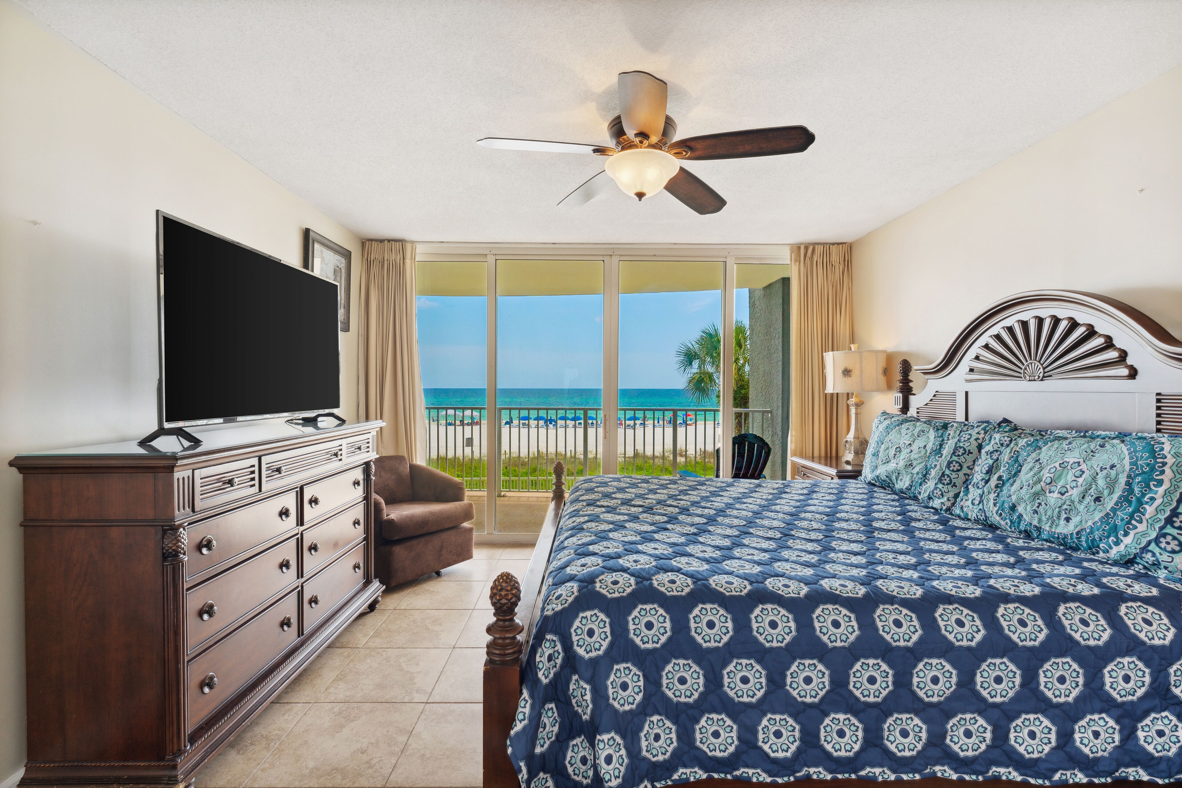 A bedroom with a large bed, a television on a dresser, and a view of the beach.