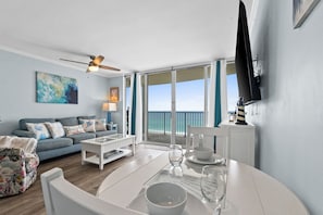 A modern living room with a sofa, coffee table, and dining table set for two. A ceiling fan and a television are mounted on the wall. - Long Beach by Book That Condo (Panama City Beach)