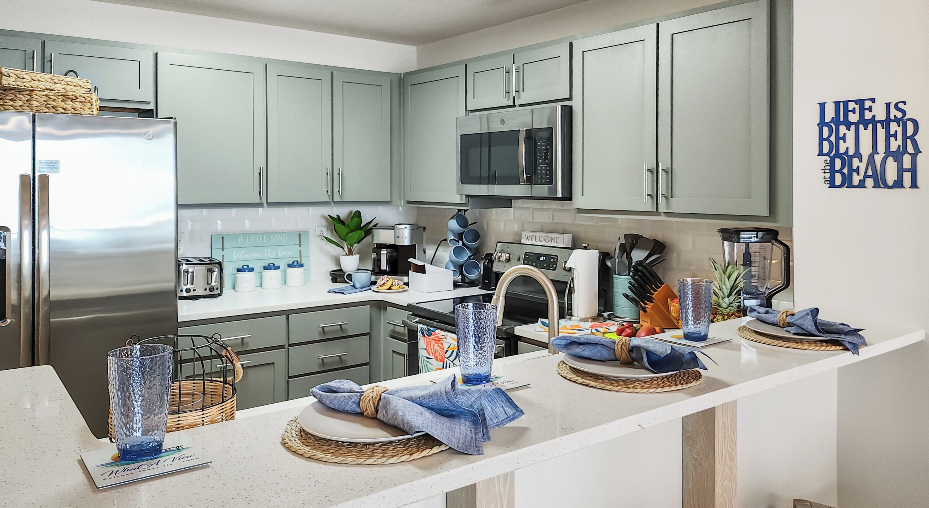 Una cucina moderna con mobili color verde chiaro, elettrodomestici in acciaio inossidabile e un piano di lavoro bianco.