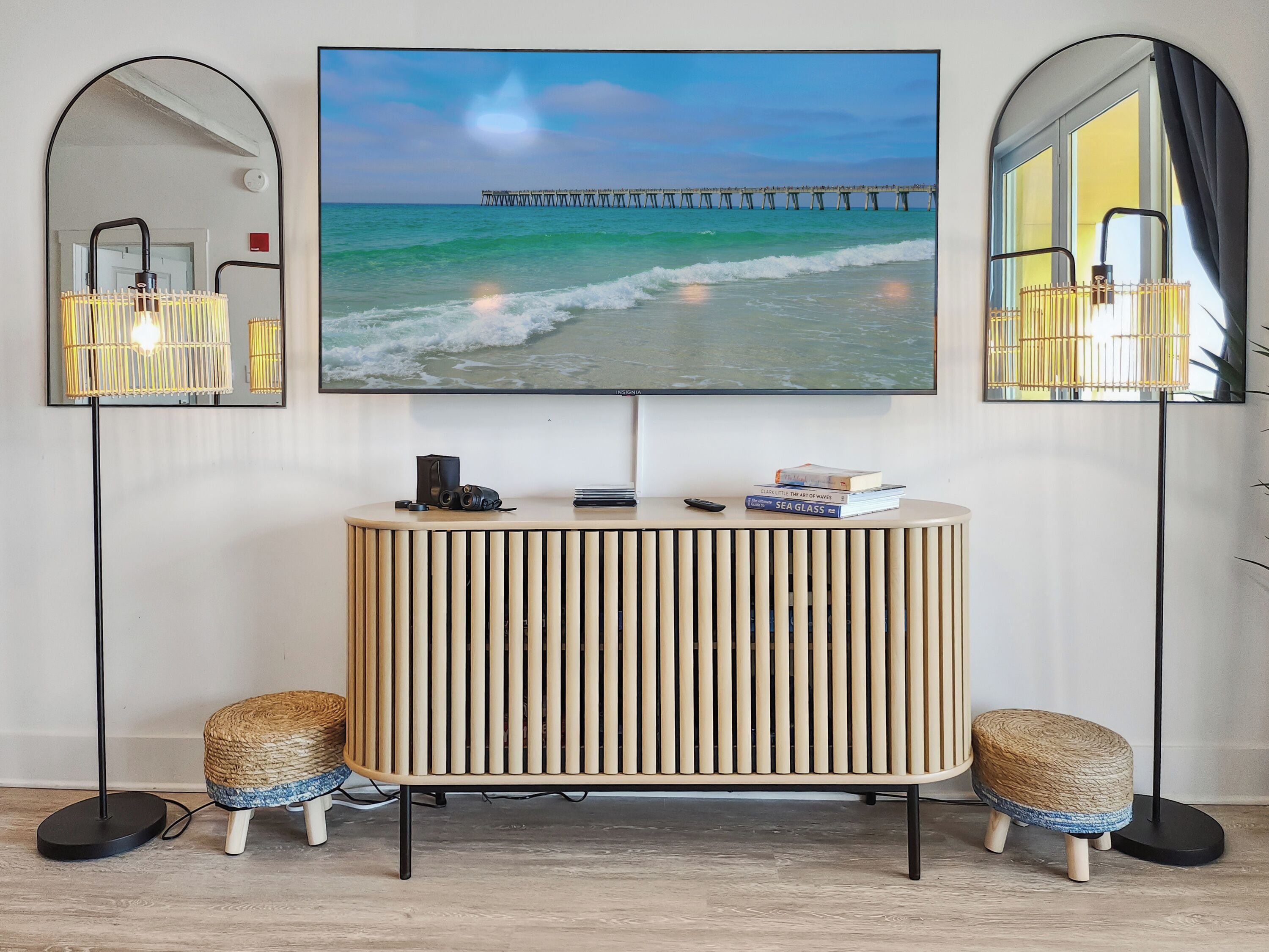 A modern living room with a large TV displaying a beach scene, a round wooden console table, two small stools, and floor lamps.