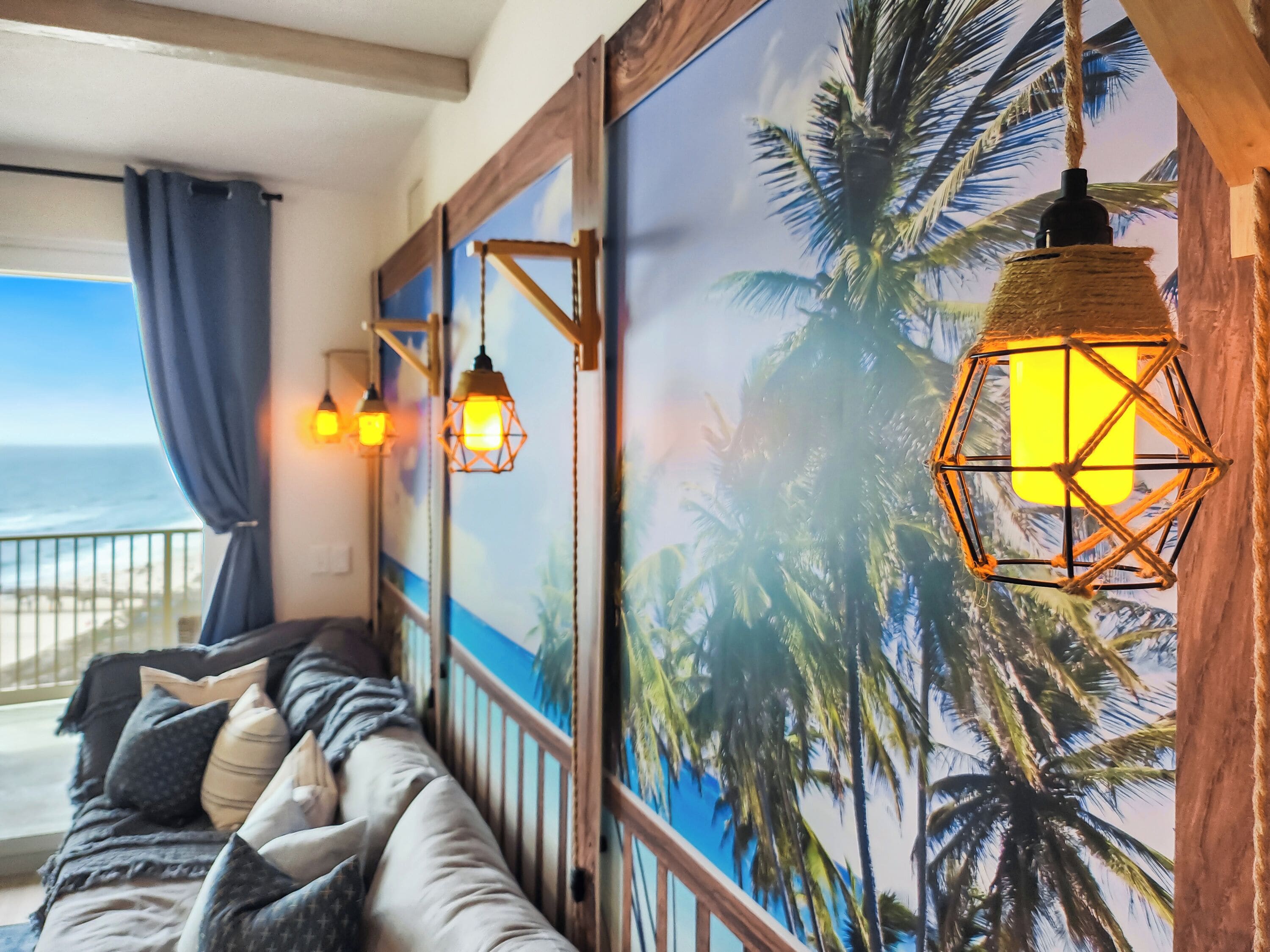 Een kamer met uitzicht op de oceaan, een houten wand met een tropische muurschildering, een comfortabele zithoek en hangende lantaarnlampen.