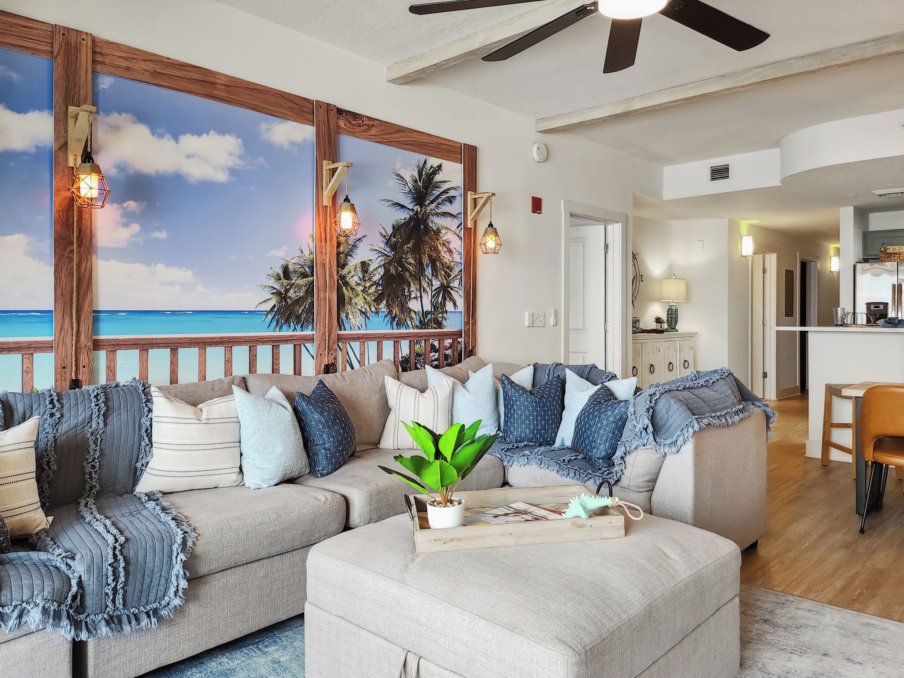 A living room with a large window overlooking a beach, a ceiling fan, and a wooden deck.