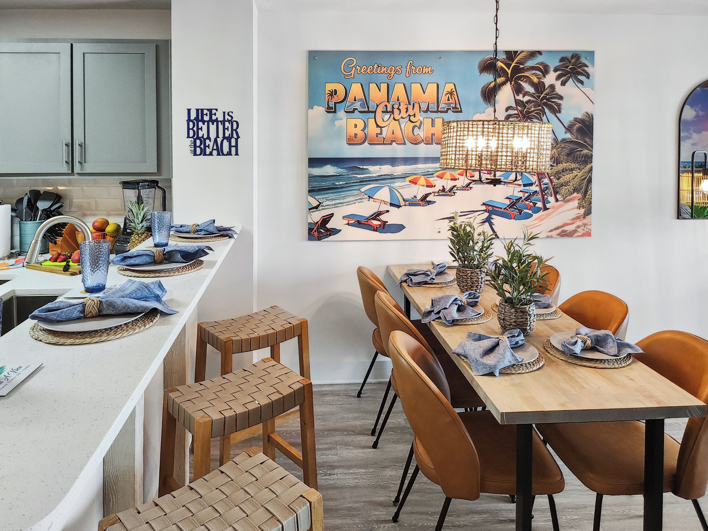 Un comedor con una larga mesa dispuesta para dos, una zona de cocina con armarios y un frutero, y una pared con un cartel que dice 'Life is Better Beach' y un póster que dice 'Greetings from Panama City Beach'.