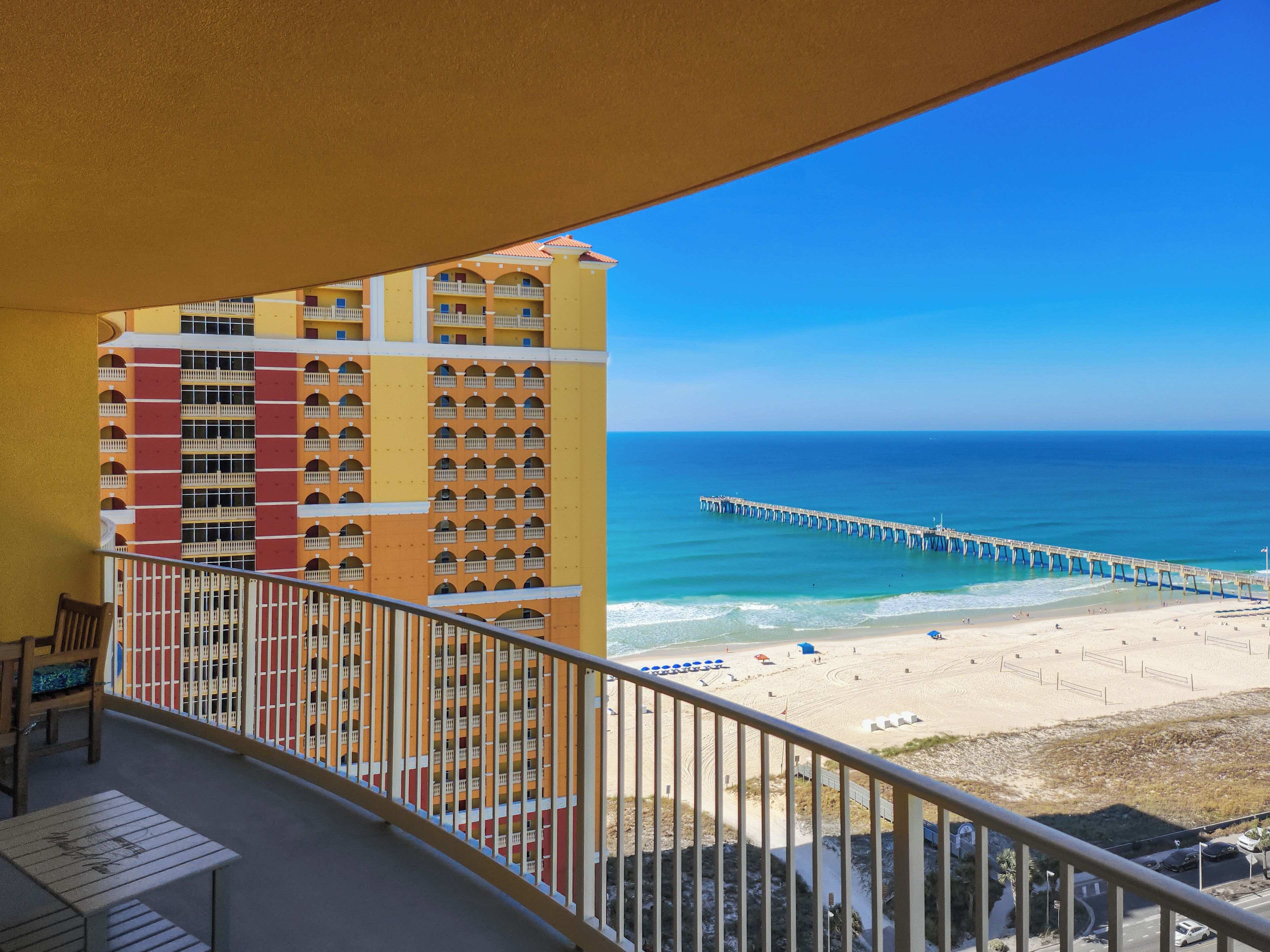 A balcony with a view of a beach, a pier, and a multi-story building with balconies.