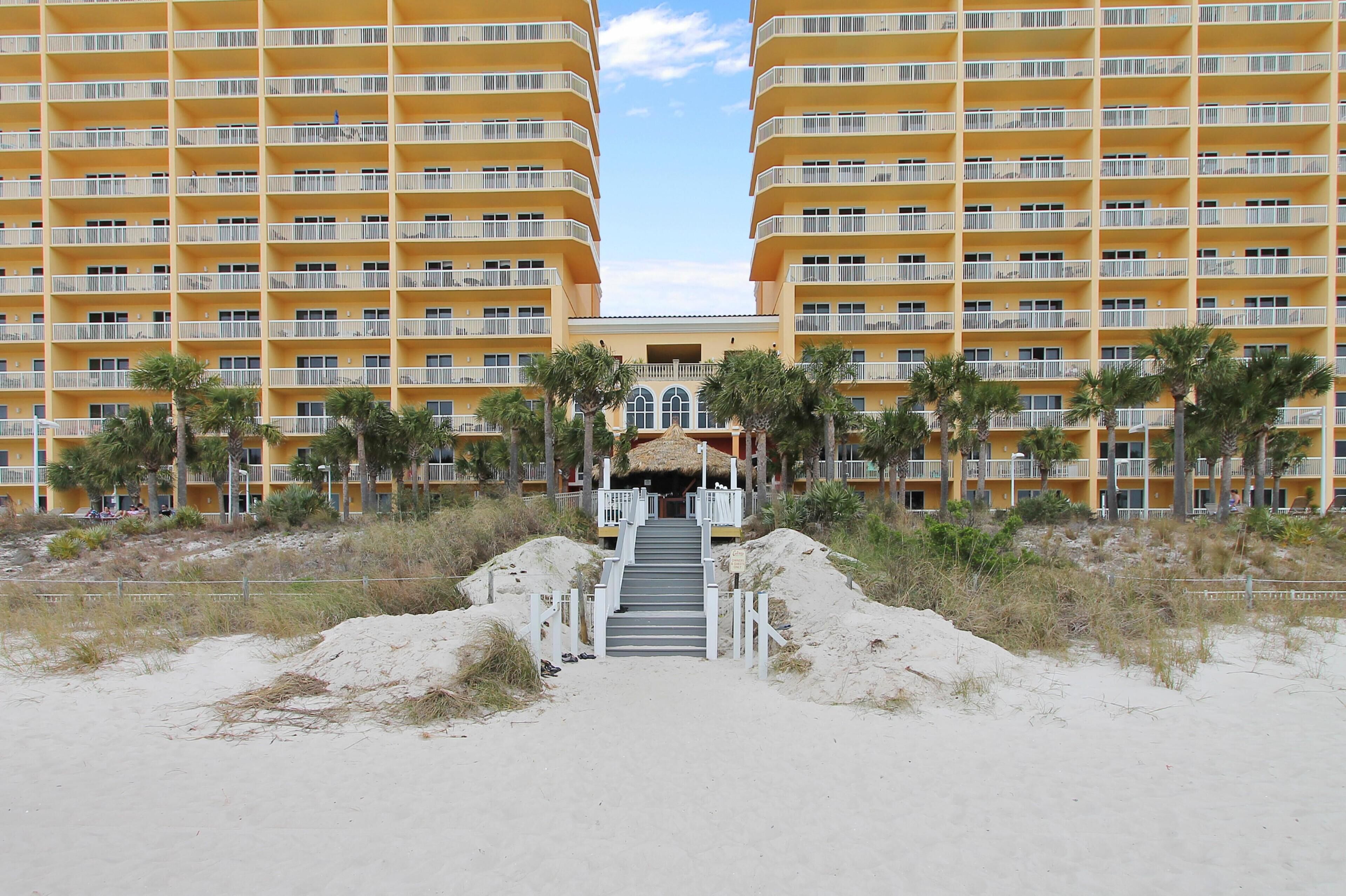Un hotel frente a la playa con una escalera central que conduce a una entrada cubierta, rodeado de palmeras y arena.