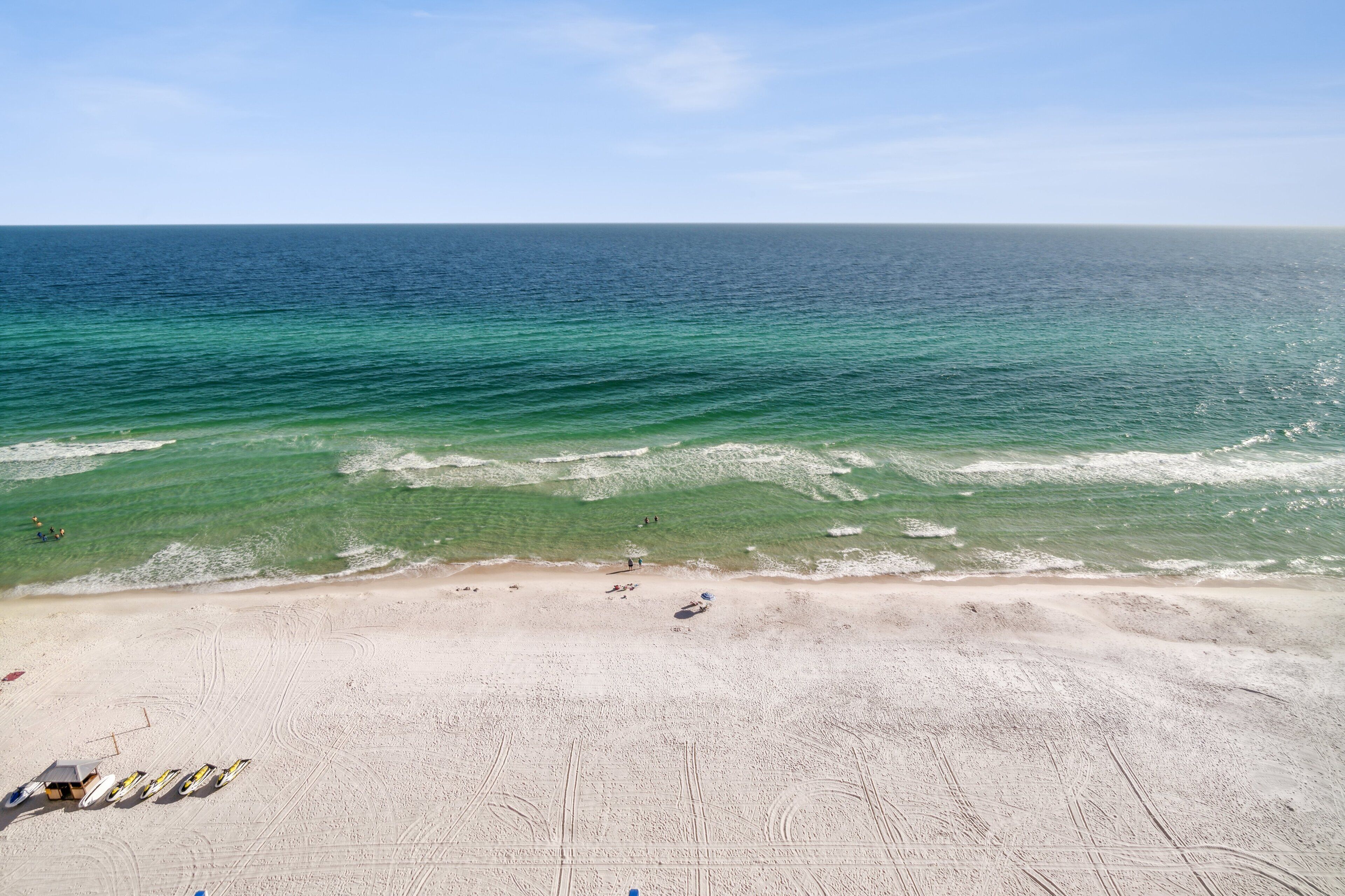 A beach with clear blue water, sandy shore, and a few people.