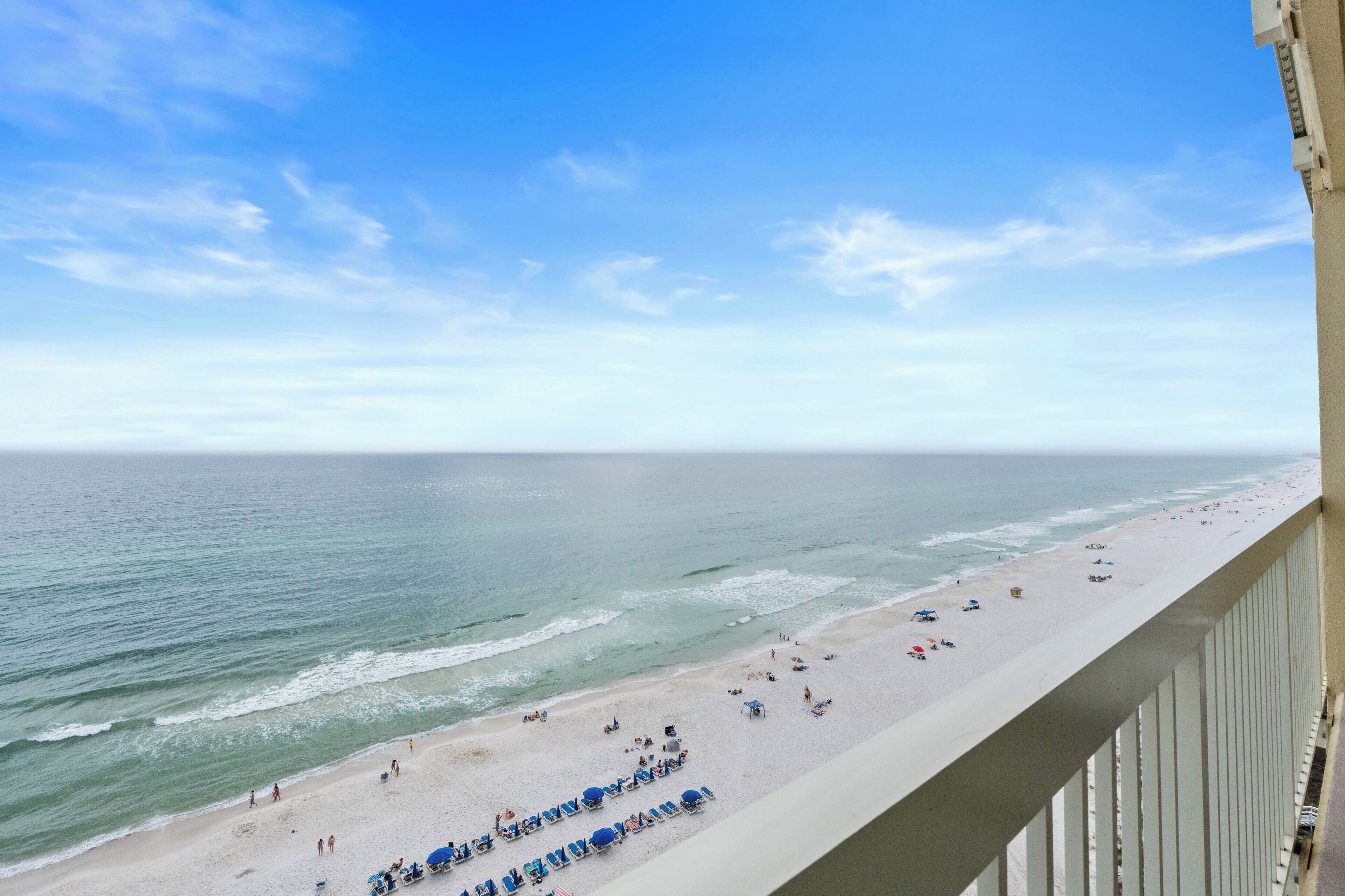 A beach view from a balcony with a railing, ocean, and beachgoers.
