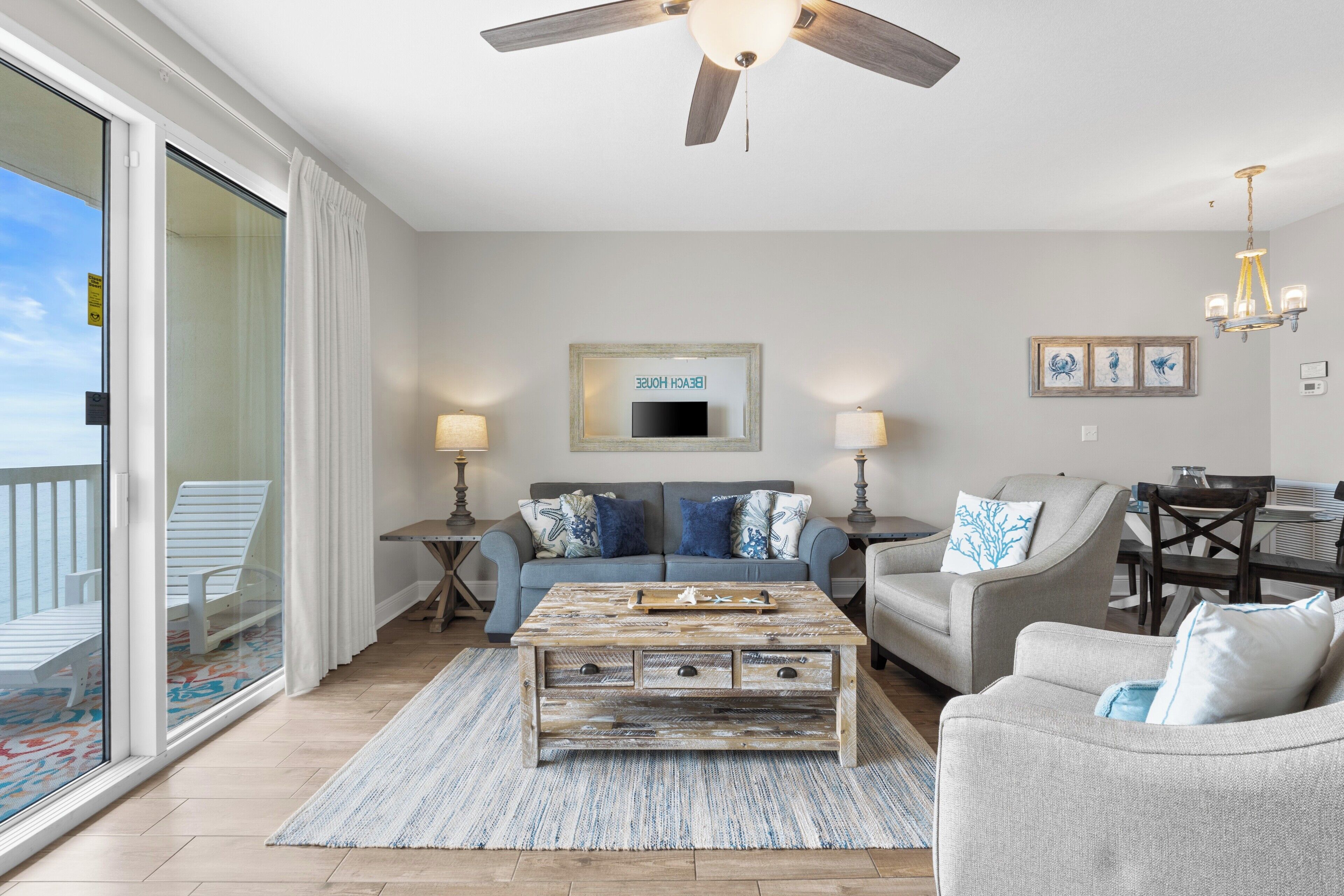 A spacious living area with a sofa, coffee table, and dining table. There is a ceiling fan, a chandelier, and a large window with a view.