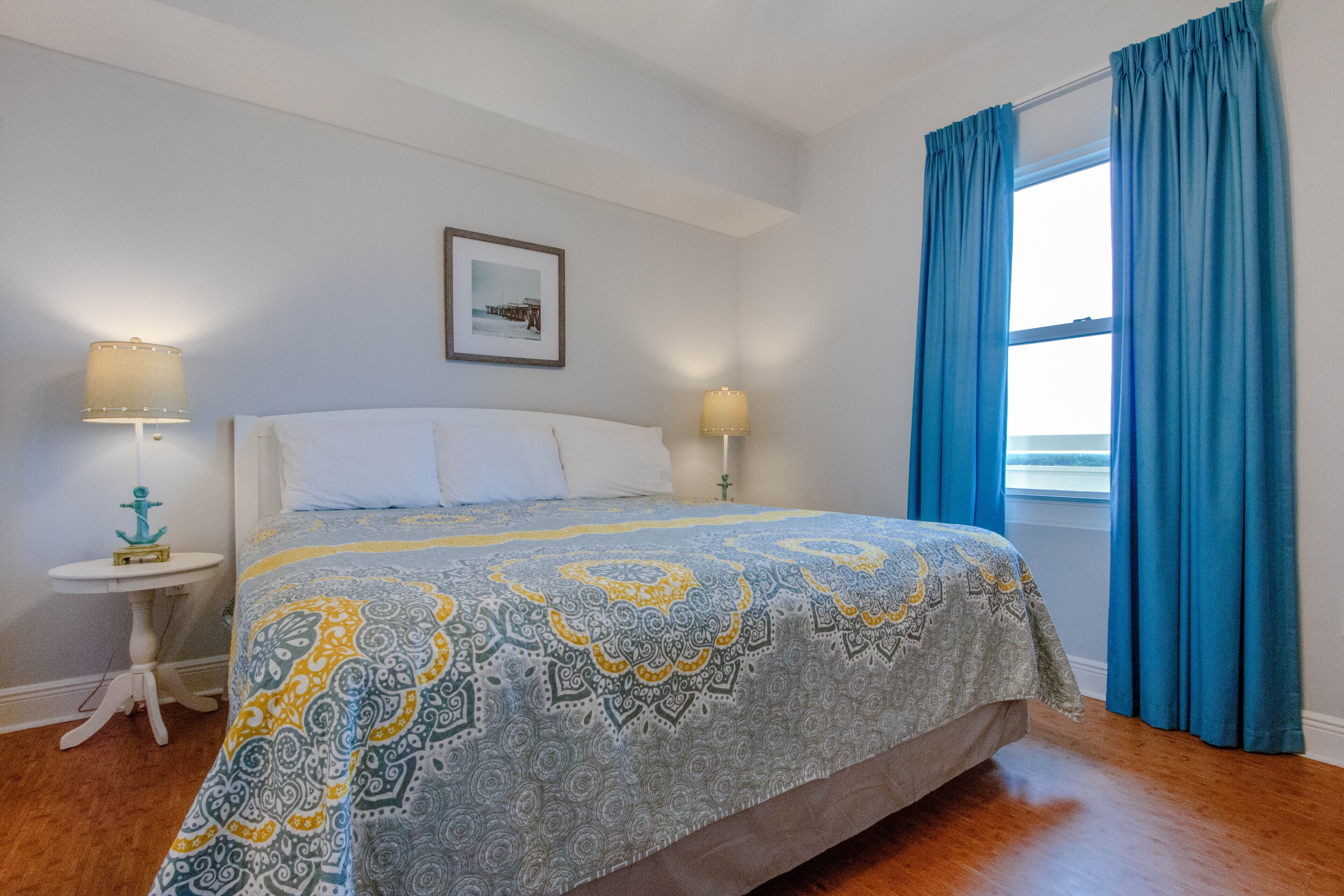 Une chambre à coucher avec un lit, deux tables de chevet, une fenêtre avec des rideaux et un tableau encadré au mur.