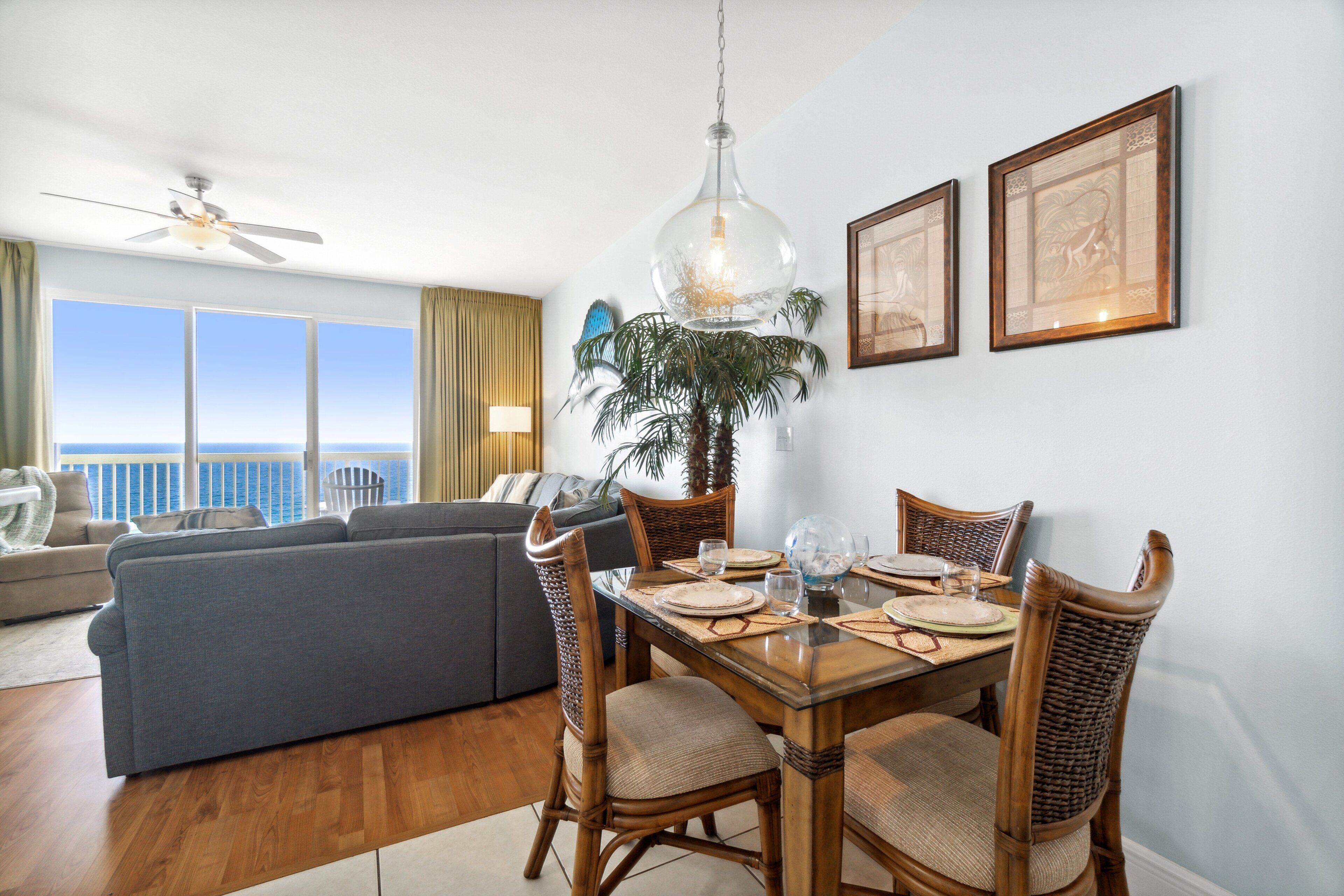 Een eetgedeelte met een houten tafel gedekt voor vier personen, omringd door rieten stoelen. Boven de tafel hangt een plafondventilator en een groot raam geeft uitzicht op de oceaan.