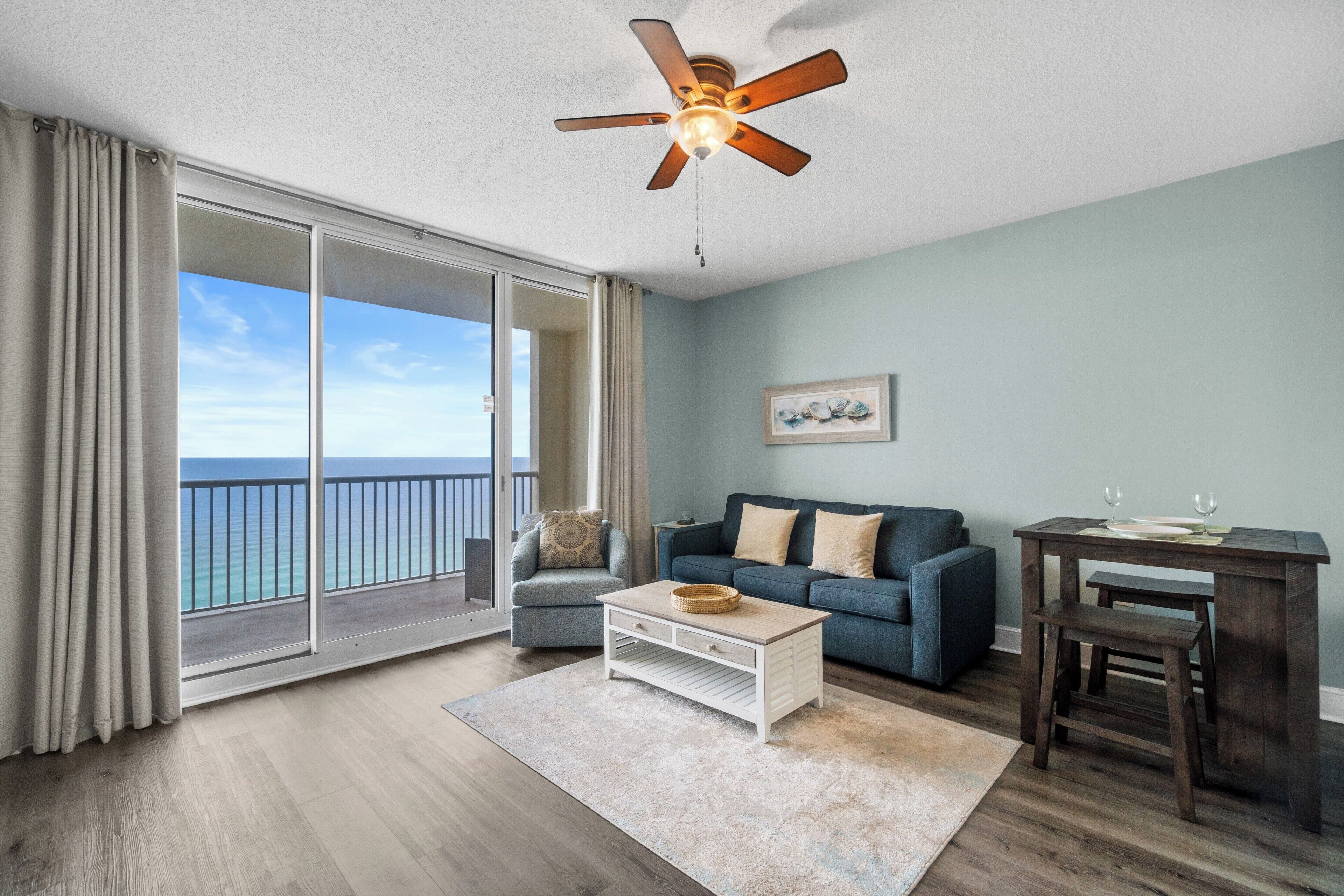 A spacious living room with a large sliding glass door leading to a balcony with a view of the ocean. There is a ceiling fan, a blue sofa, a coffee table, and a small wooden table with a bench.