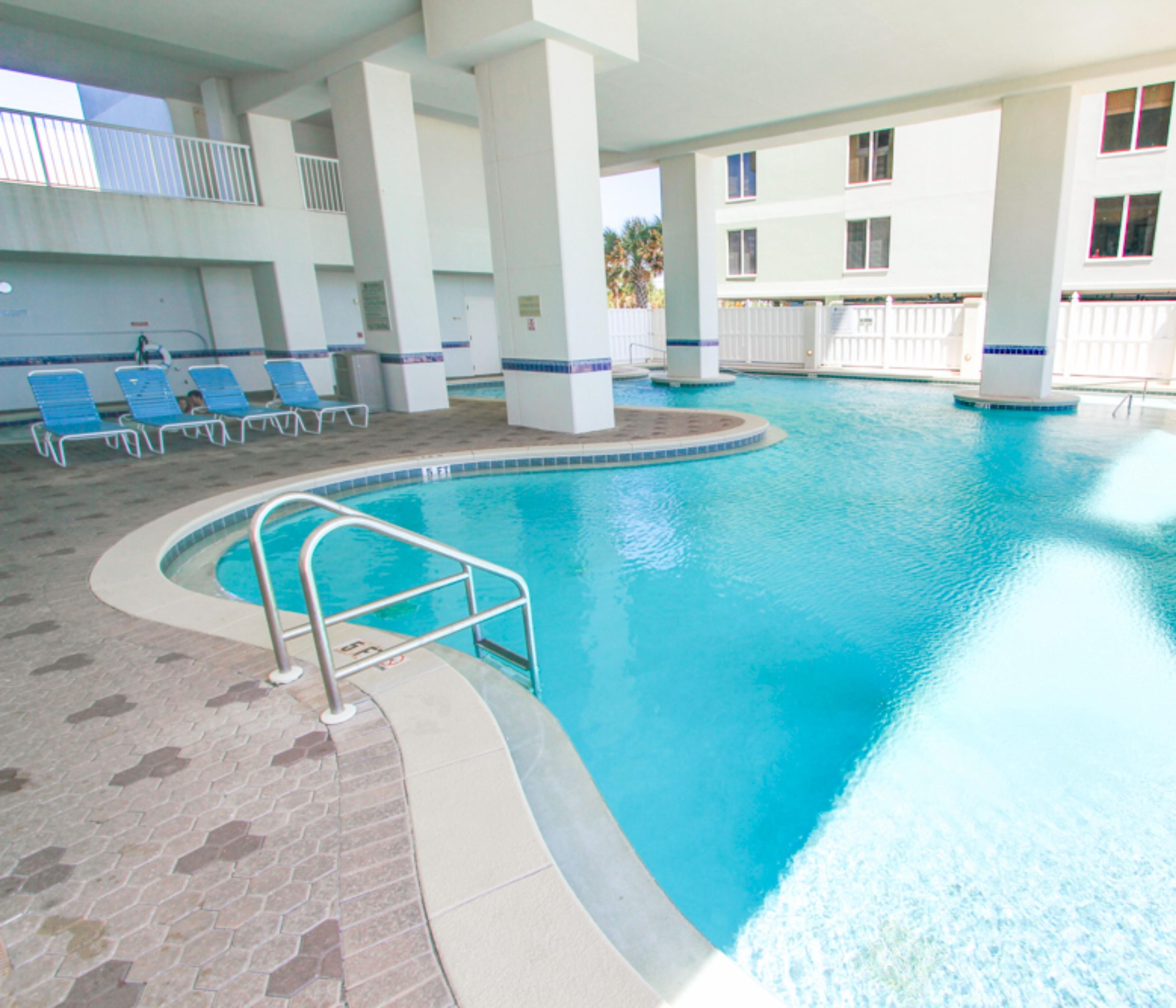 An indoor swimming pool with a tiled floor, a pool ladder, and blue lounge chairs.