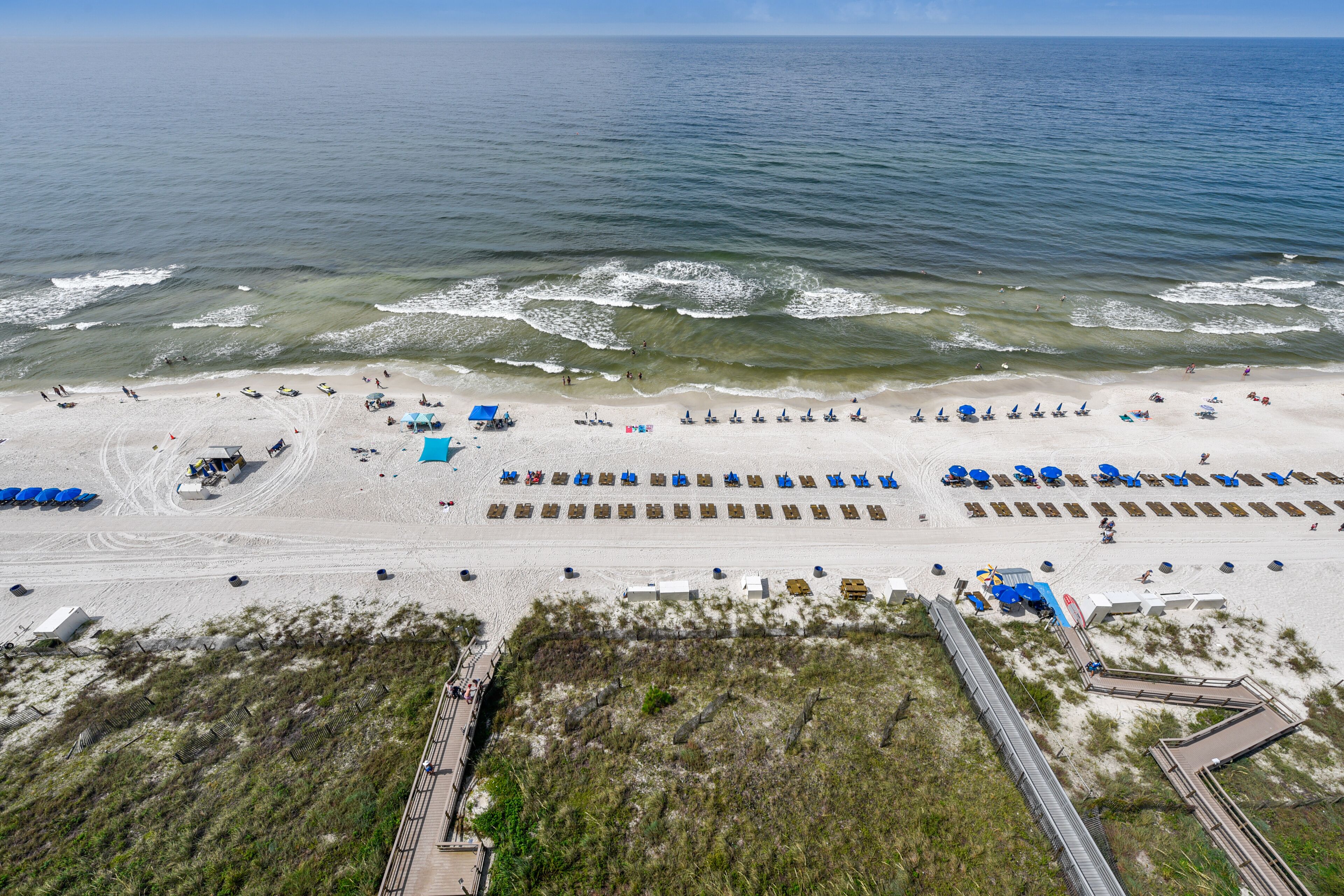 Vista aérea de una playa con hileras de sombrillas azules, arena y un paseo marítimo que conduce al agua.