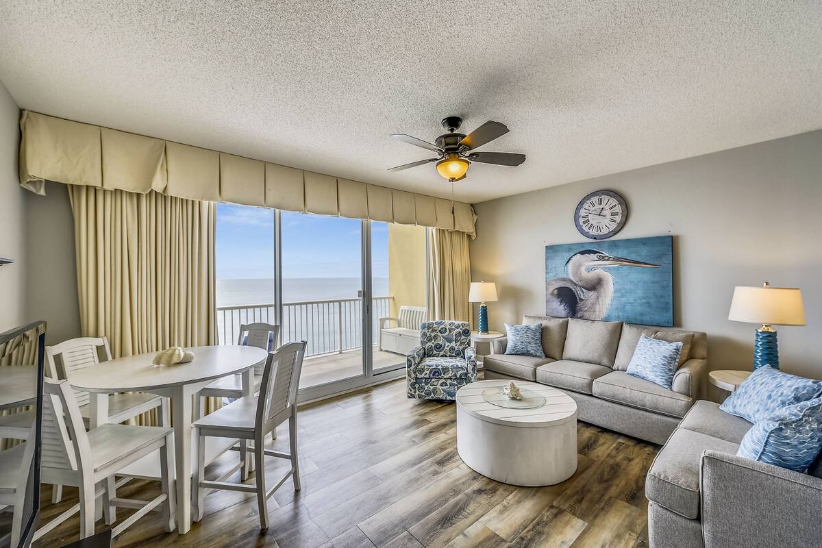 A spacious living room with a balcony, a ceiling fan, a round coffee table, and a painting of a bird.