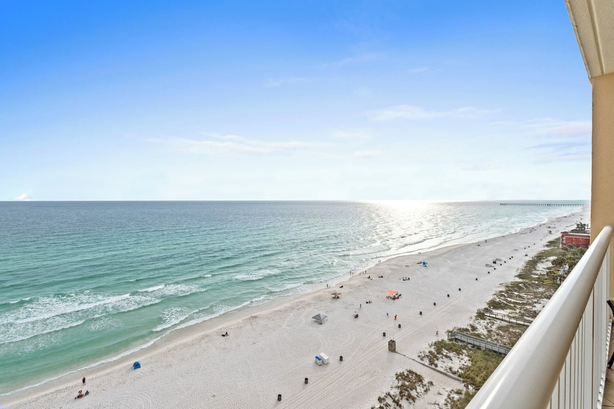A beach view from a balcony overlooking a sandy beach with a pier in the distance.