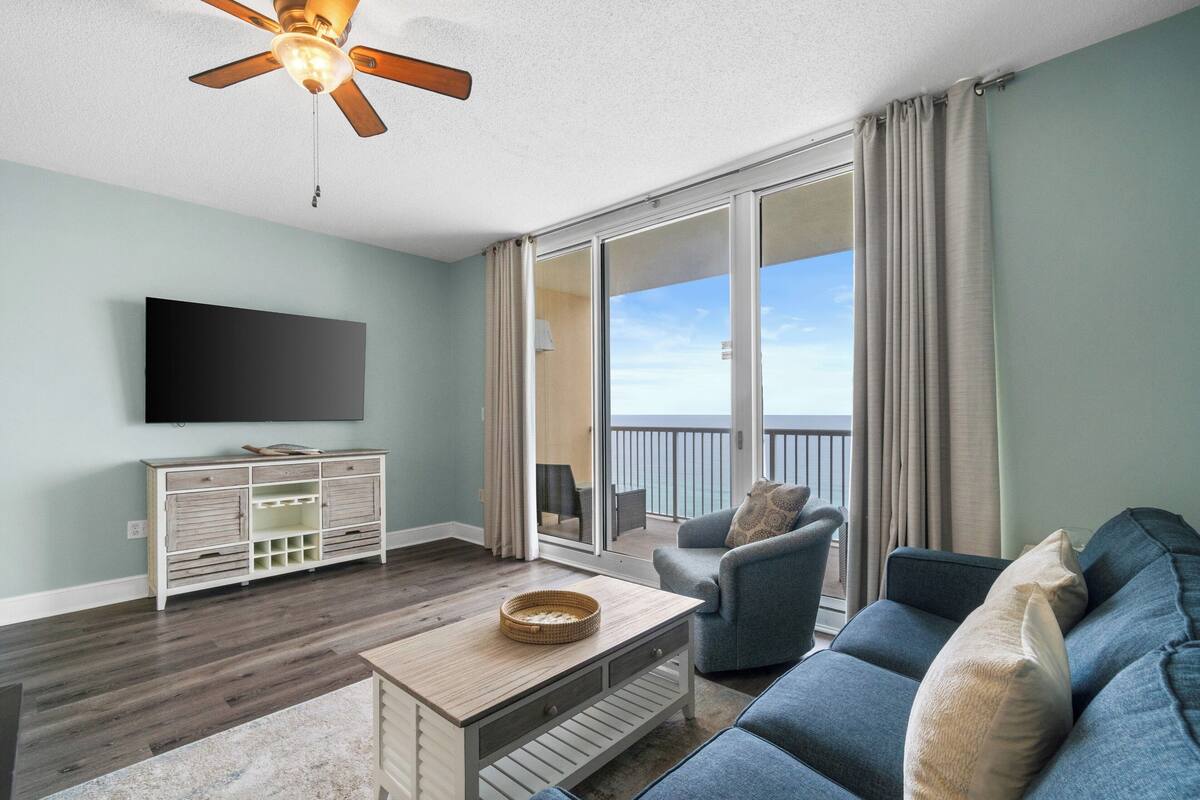 A living room with a flat-screen TV, a wooden coffee table, a blue sofa, and a ceiling fan.