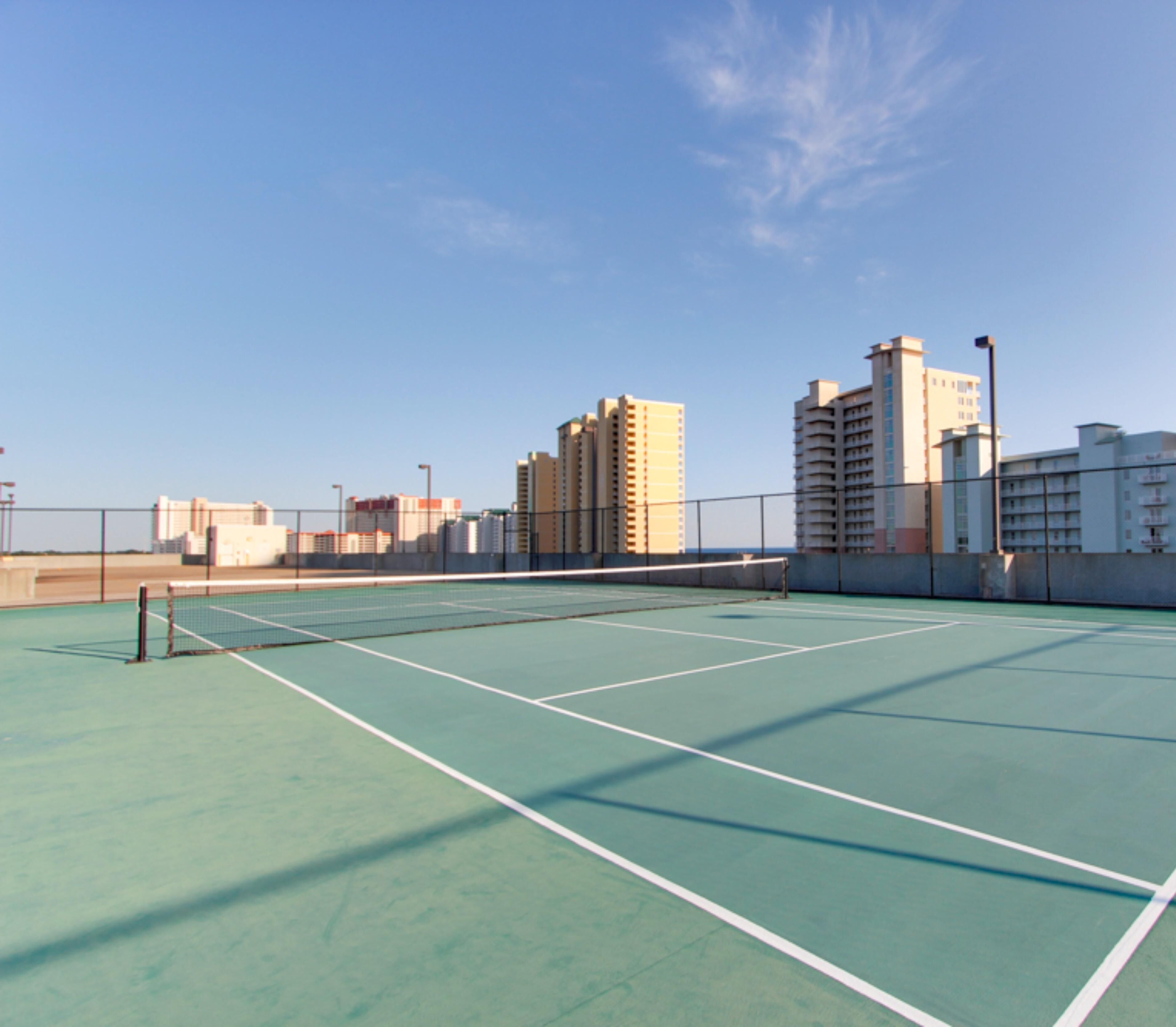 Una cancha de tenis al aire libre con cielo despejado, rodeada de edificios altos.