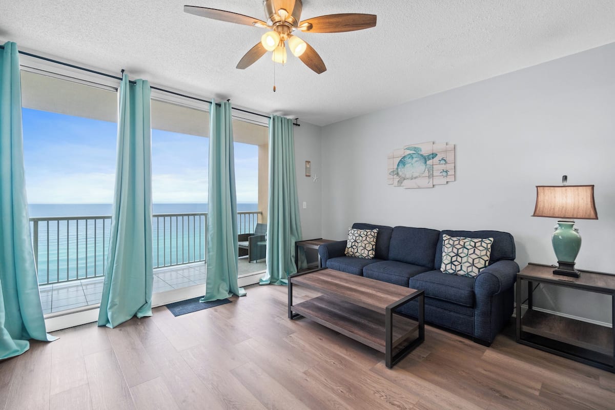 A living room with a sofa, coffee table, and lamp, a ceiling fan, and a view of the ocean through large windows.