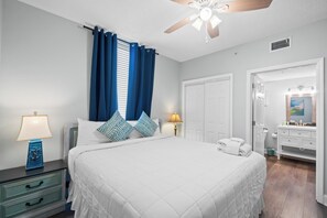 A bedroom with a bed, nightstands, a ceiling fan, a window with curtains, a door, and a bathroom with a sink and mirror. - Majestic Beach Resort by Book That Condo (Panama City Beach)