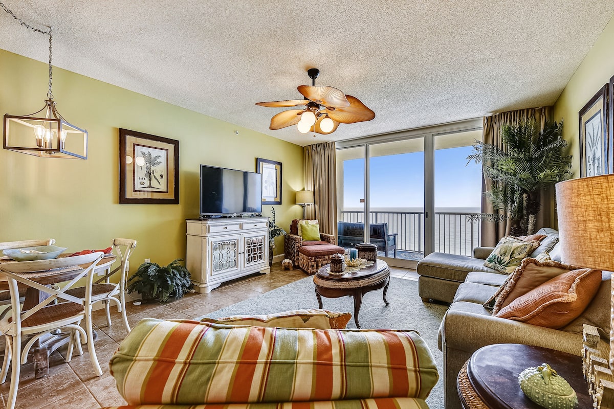A spacious living room with a dining area, a ceiling fan, a television, and a large window offering a view of the outdoors.