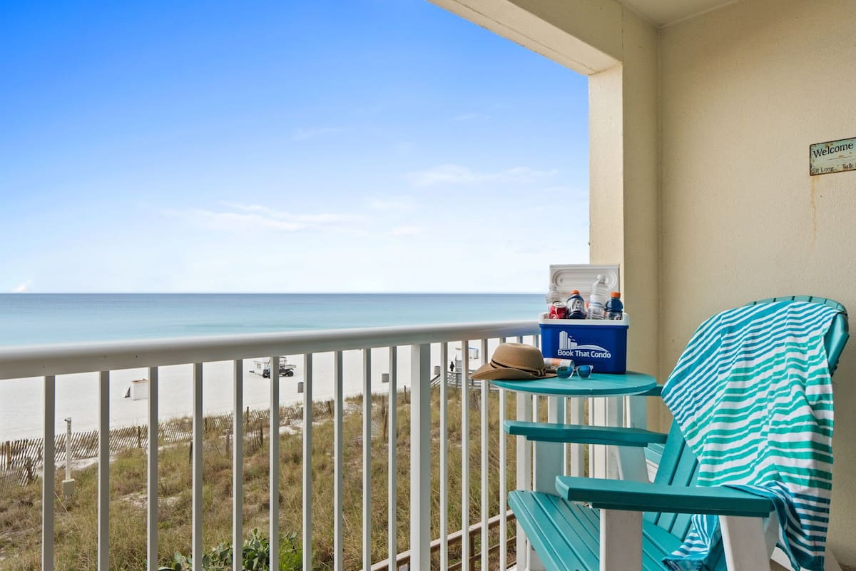 A balcony with a view of the ocean, a blue cooler with drinks, and a straw hat on a chair.