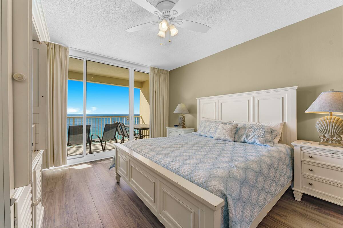 A bedroom with a large bed, a nightstand, a lamp, and a view of the ocean through a sliding glass door.