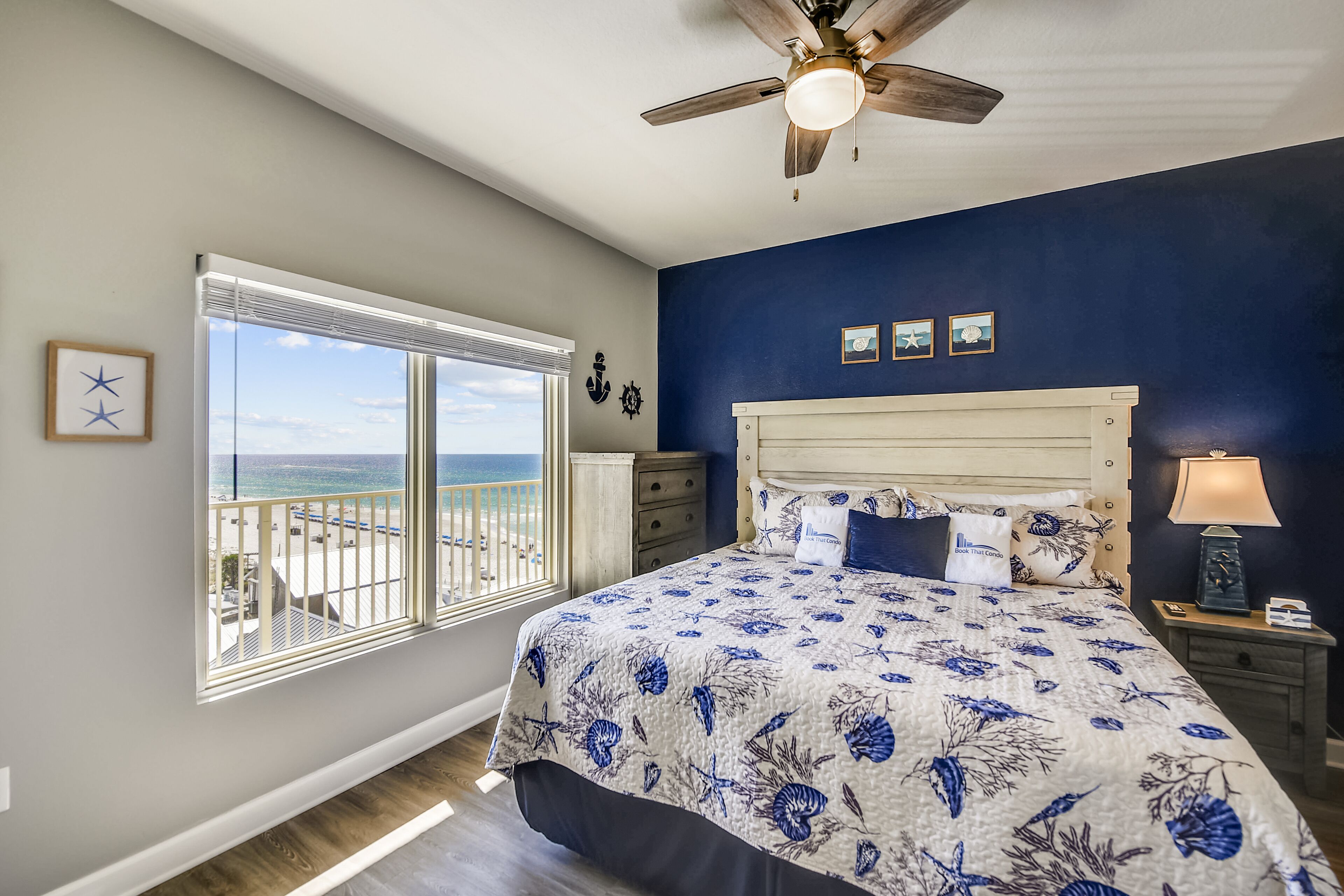 A bedroom with a large bed, a ceiling fan, a window with a view of the beach, and a nightstand with a lamp.