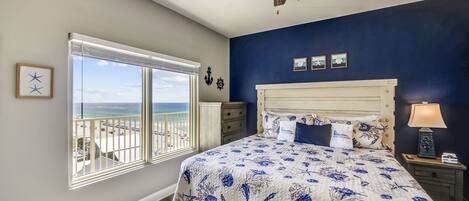 A bedroom with a large bed, a ceiling fan, a window with a view of the beach, and a nightstand with a lamp.