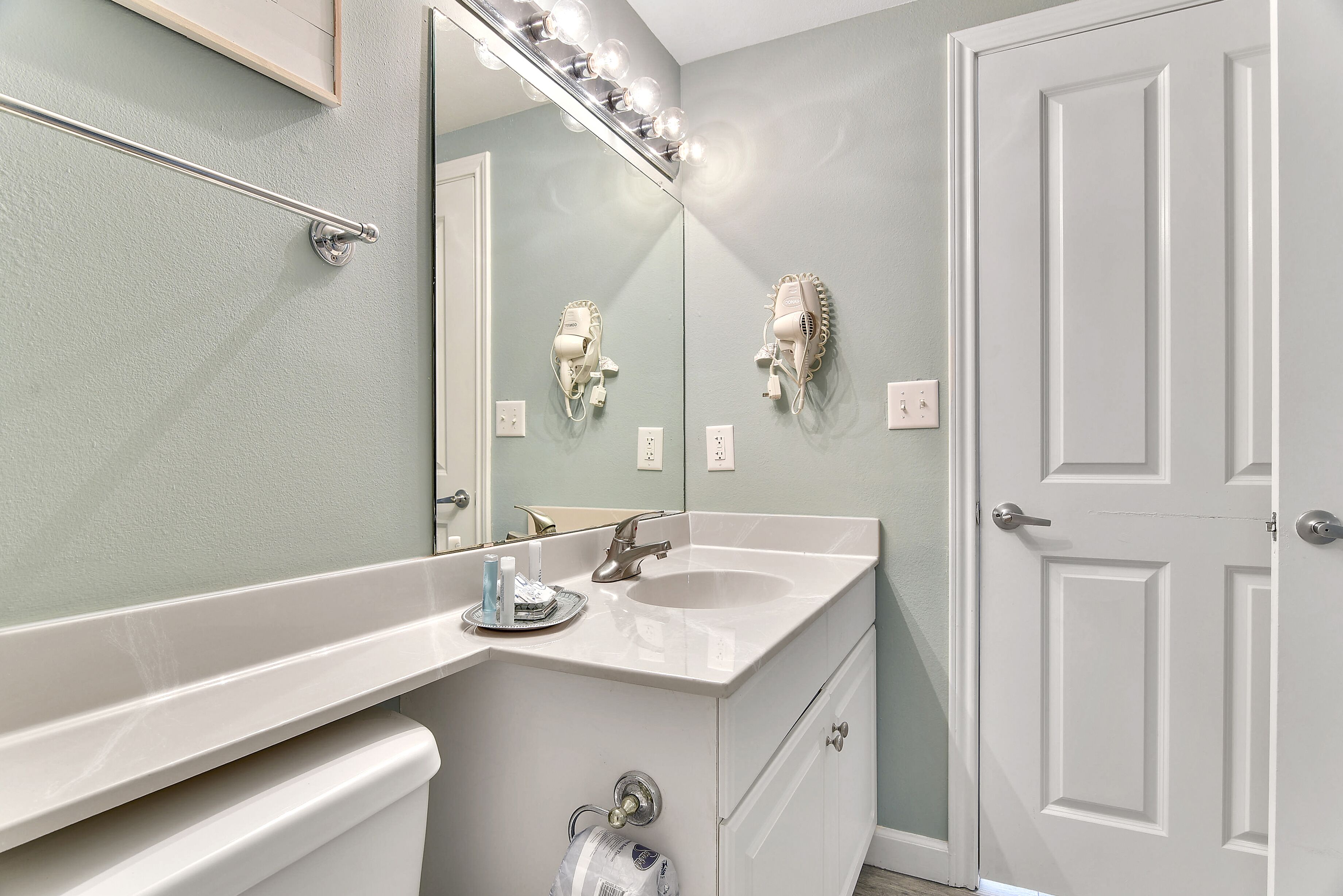 A bathroom with a white sink, a mirror with lights, a towel rack, and a door.