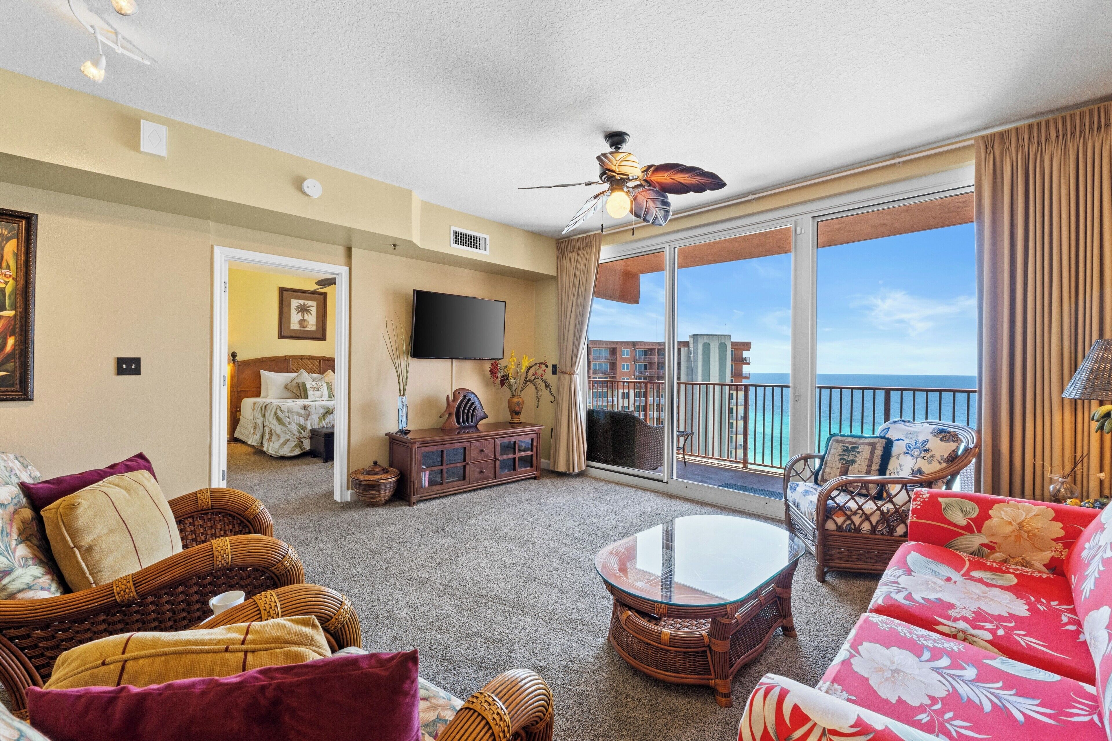 A spacious living room with a large window, a ceiling fan, and a balcony with a view of the ocean.