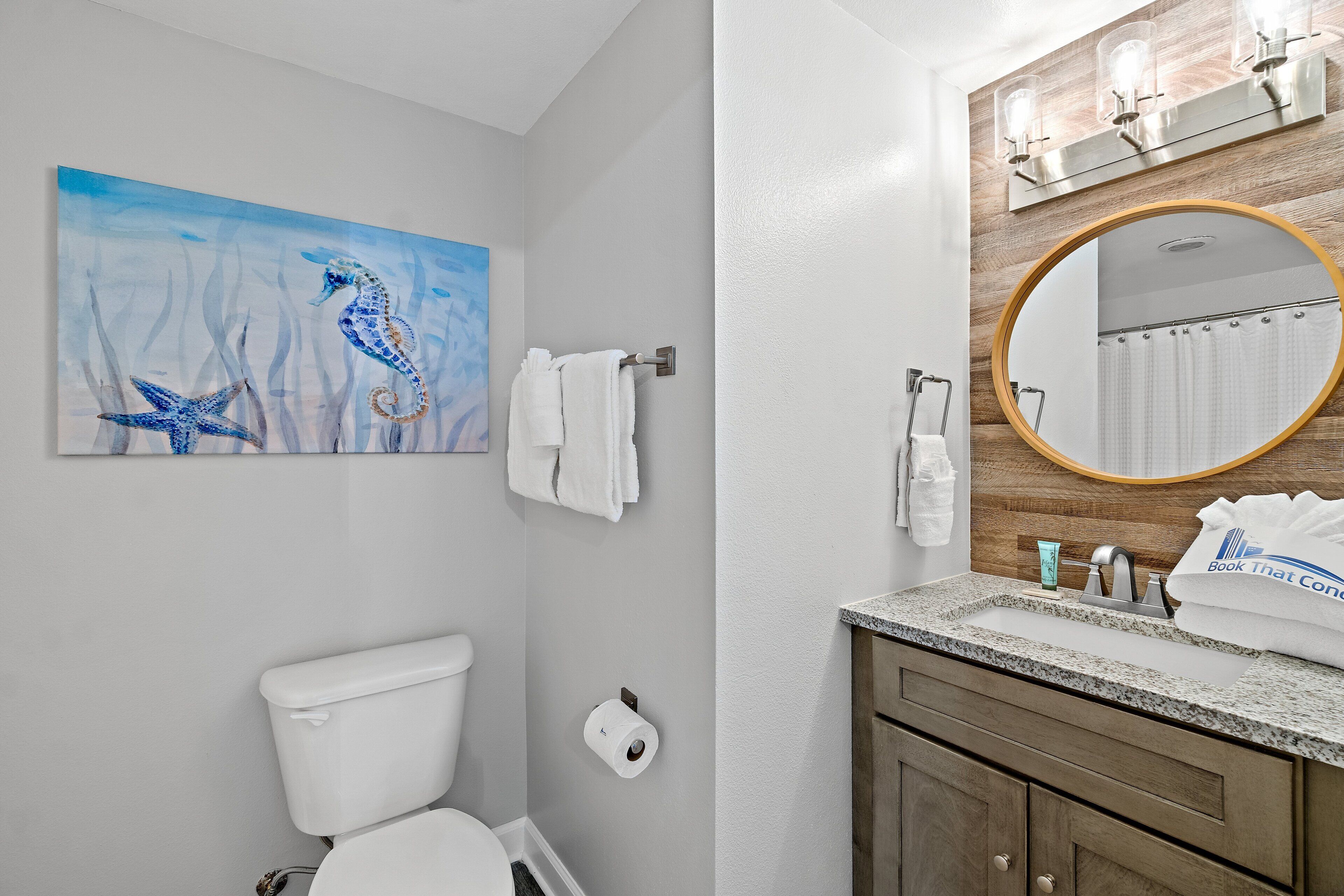 A bathroom with a toilet, a towel rack, a painting of seahorses, a sink with a mirror, and a towel with a hotel logo.