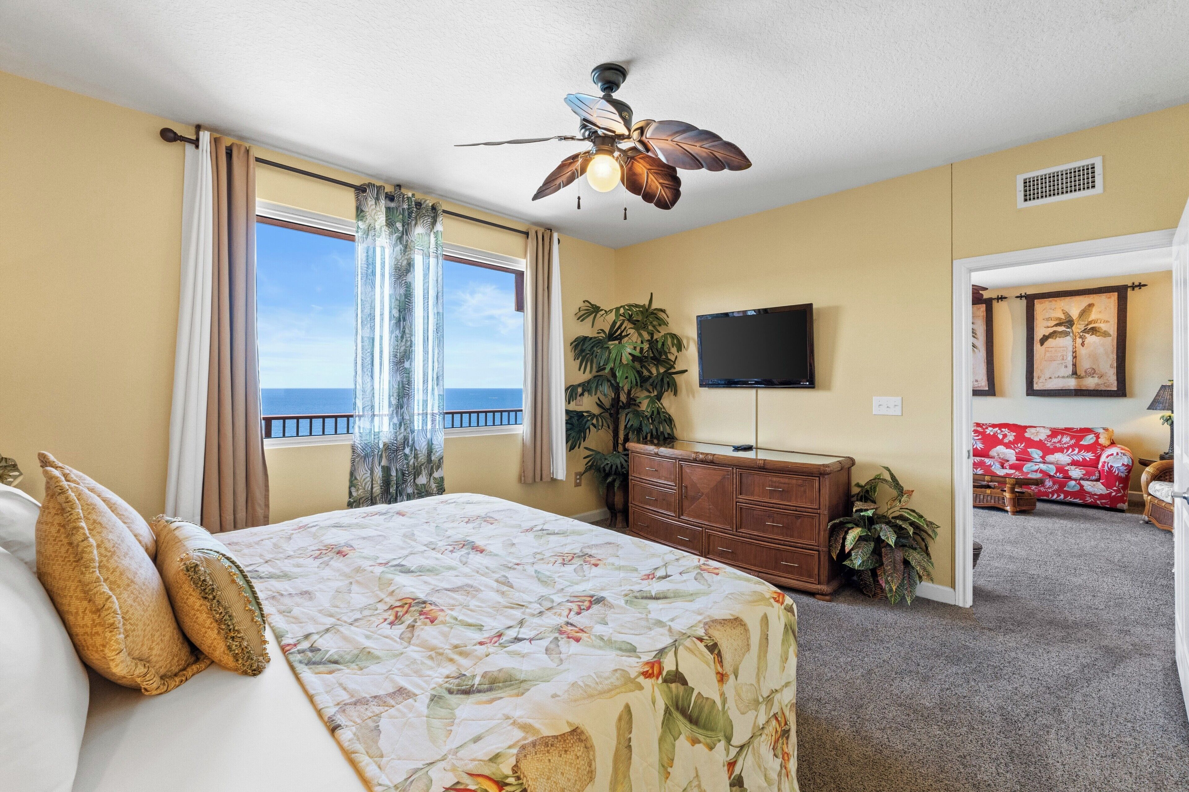 A bedroom with a bed, a ceiling fan, a TV, a plant, and a view of the ocean.