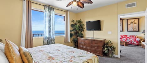 A bedroom with a bed, a ceiling fan, a TV, a plant, and a view of the ocean.