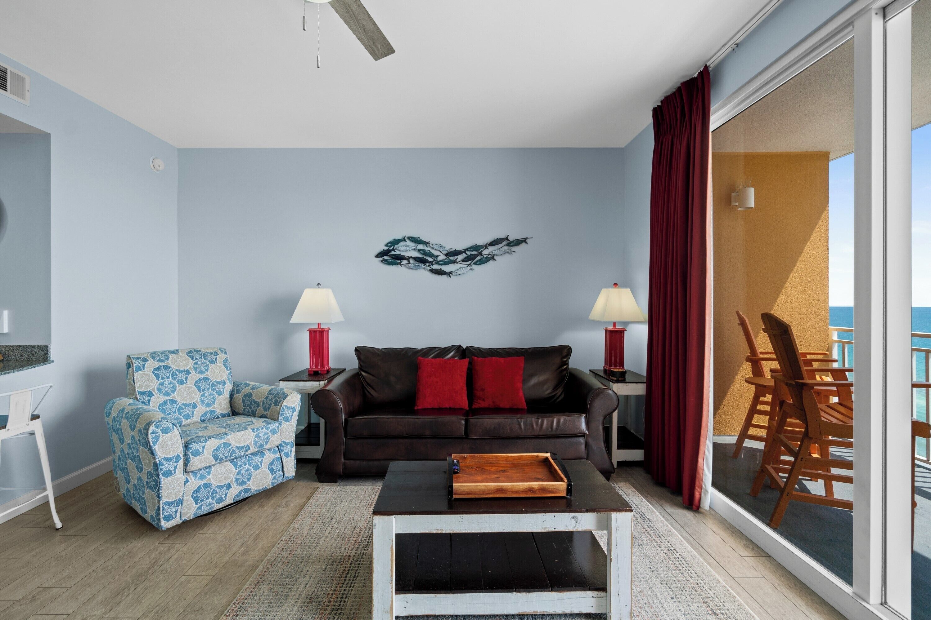A living room with a blue and white patterned armchair, a brown leather sofa with red cushions, a wooden coffee table, and a wall art piece.