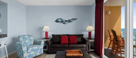 A living room with a blue and white patterned armchair, a brown leather sofa with red cushions, a wooden coffee table, and a wall art piece.