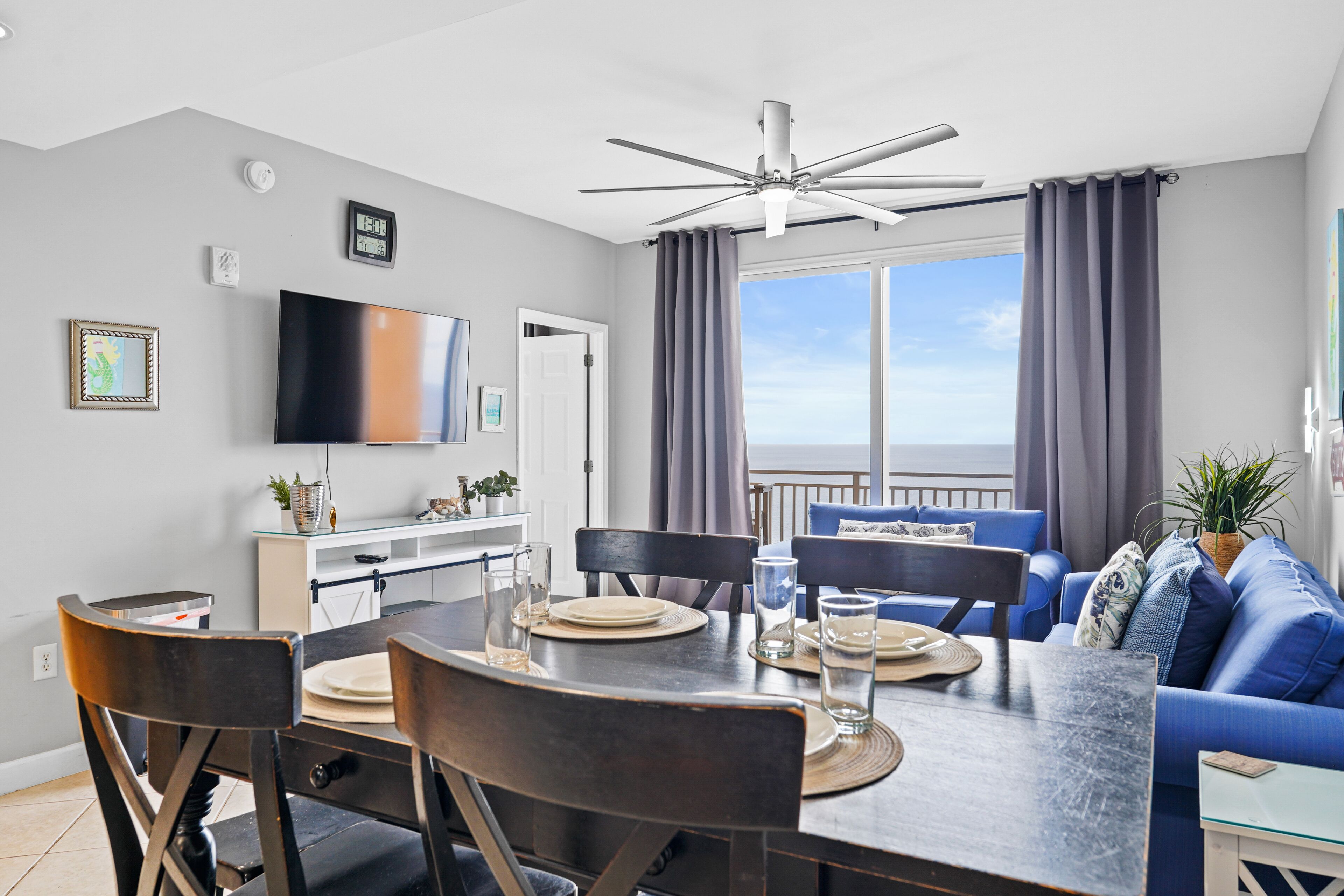 A modern living room with a dining area, a ceiling fan, a large TV, and a view to the outside.