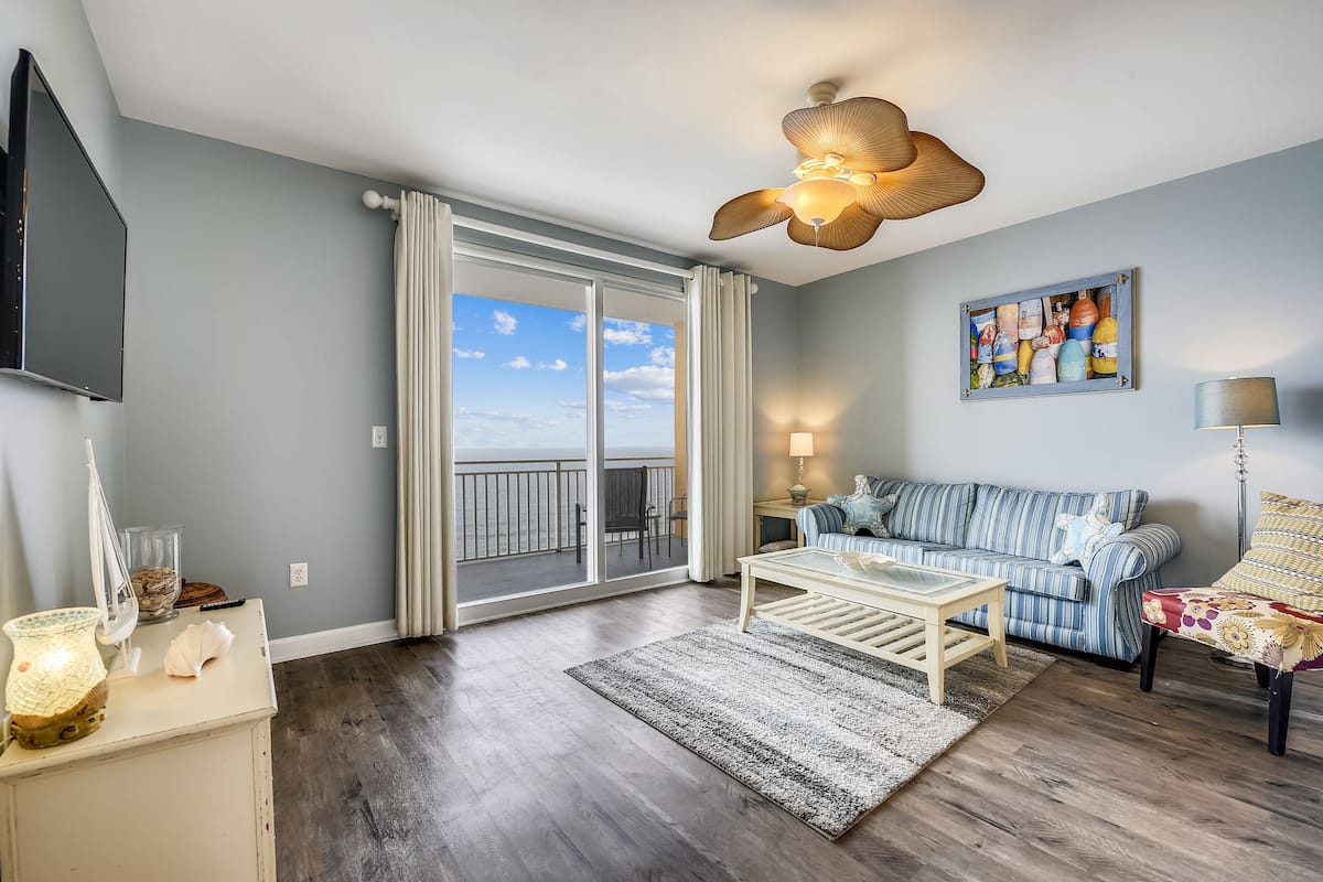 A living room with a large sliding glass door to the balcony, a flat-screen TV, a sofa, a coffee table, and a ceiling fan.