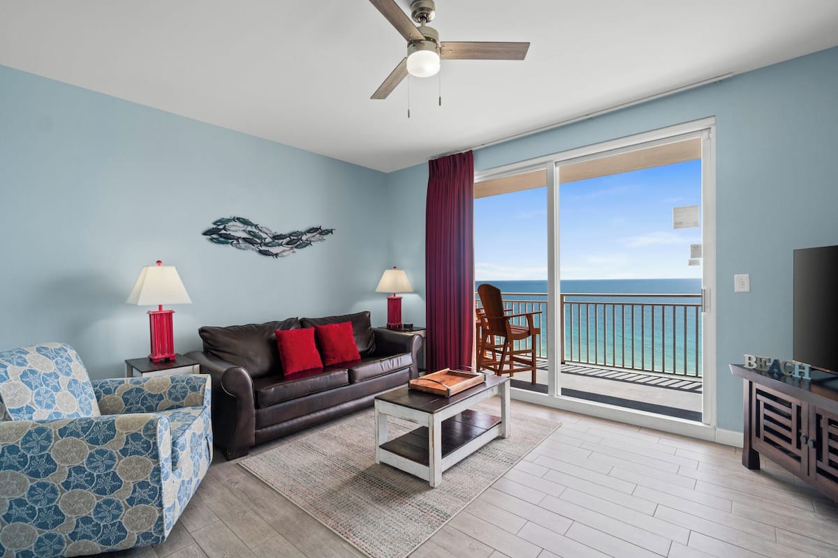 A living room with a sofa, coffee table, and chairs, a ceiling fan, and a view of the ocean through a sliding door.