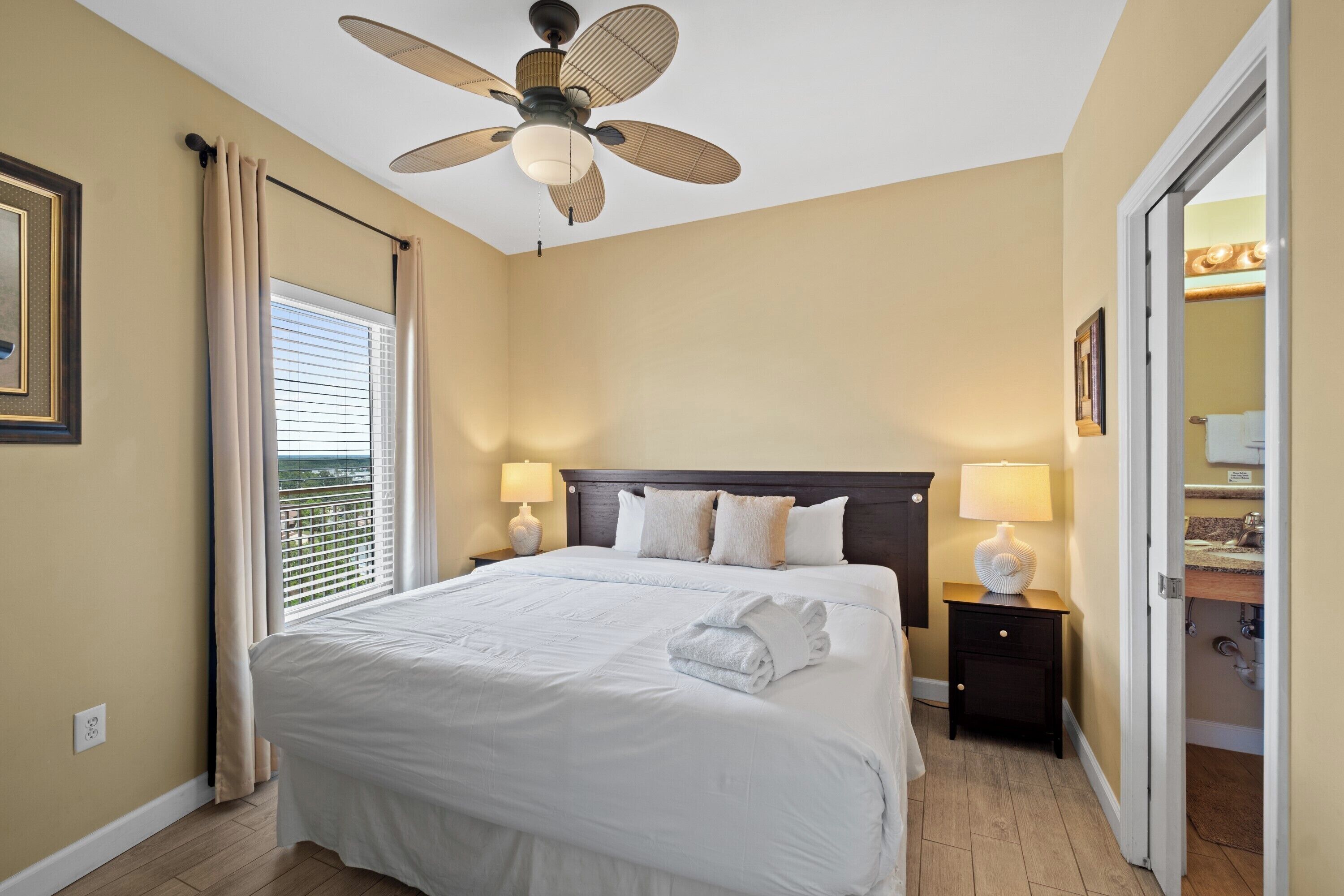 A hotel room with a large bed, two bedside tables with lamps, a ceiling fan, and a view of the outdoors through the window.