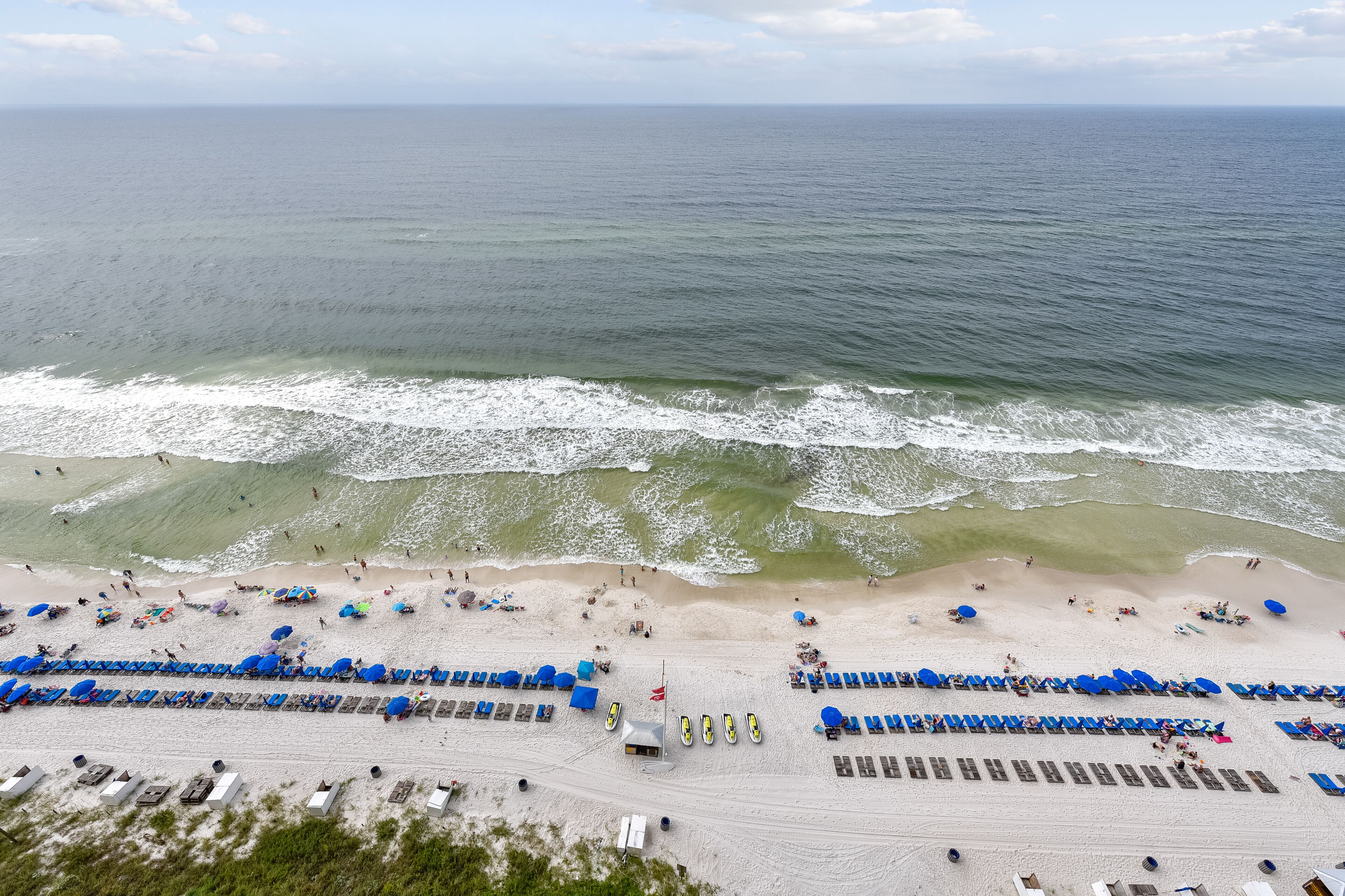 An aerial view of a beach with rows of blue lounge chairs and umbrellas, a sandy beach, and the ocean in the background.