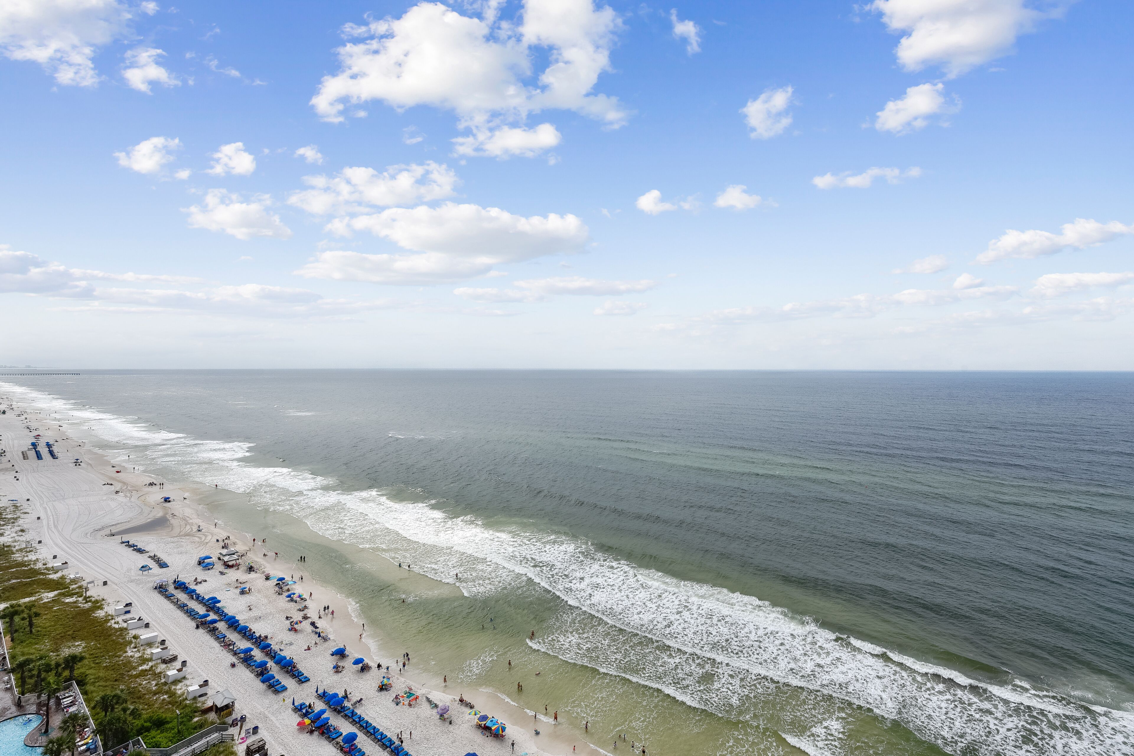 Ein Strand mit Reihen blauer Sonnenschirme und Liegestühle, ein klarer Himmel und das Meer.