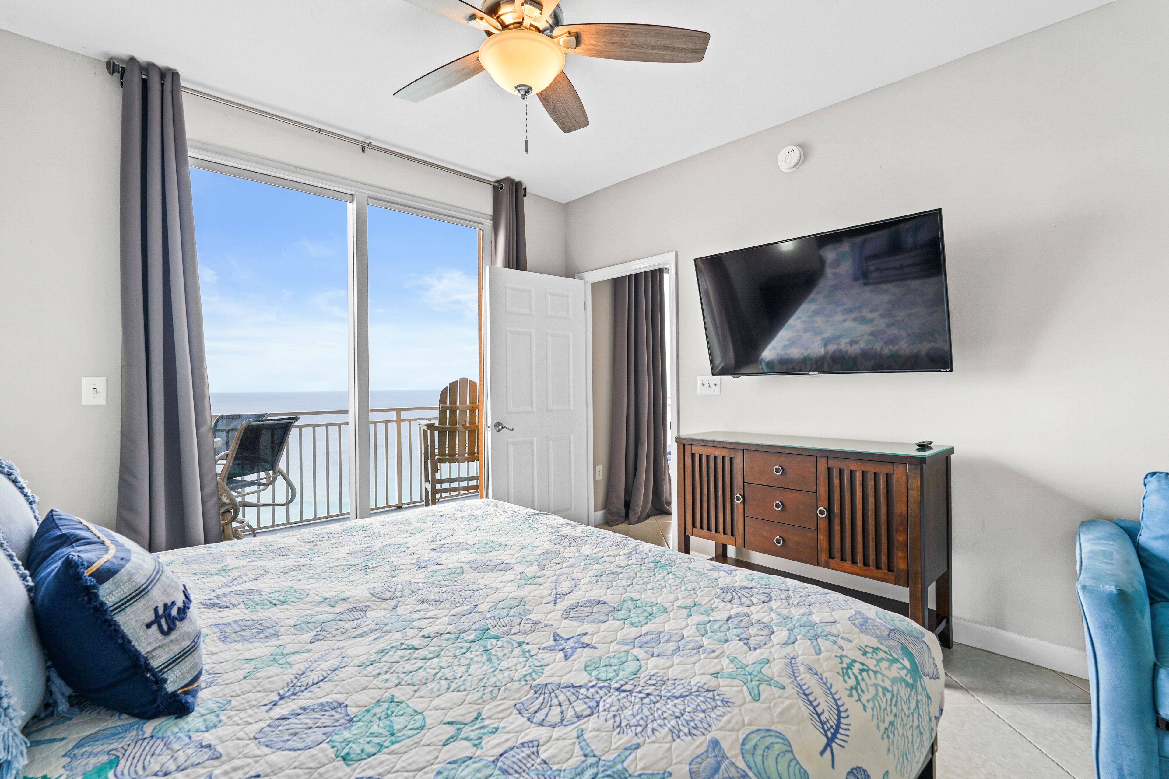 A bedroom with a bed, a ceiling fan, a TV, a wooden dresser, and a balcony with a chair.