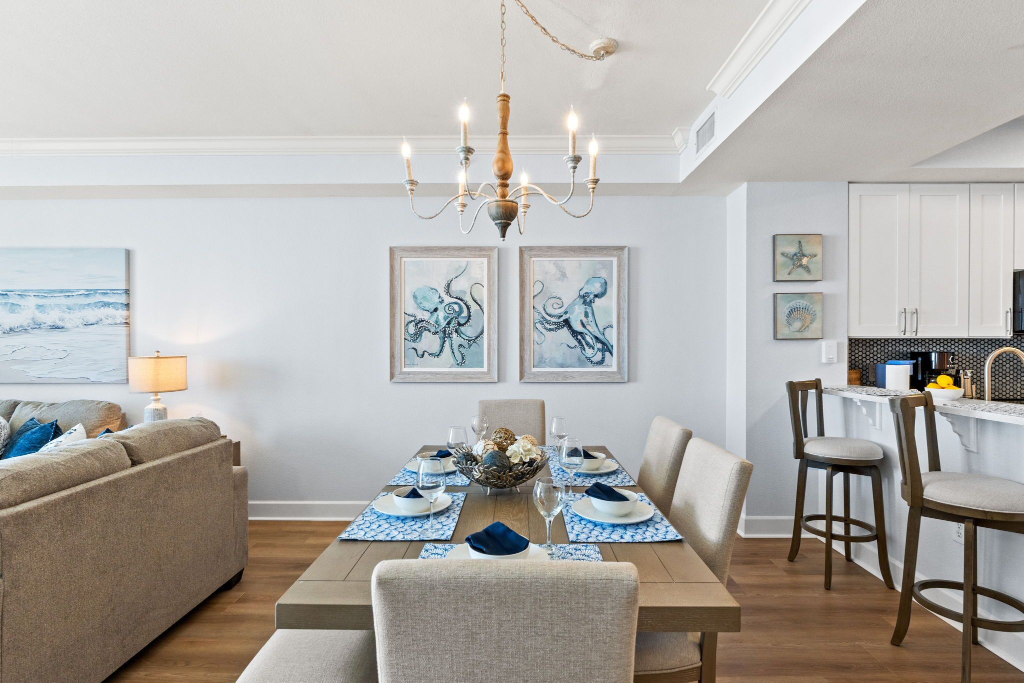 Un coin repas avec une table en bois dressée pour quatre personnes, entourée de chaises, d’un lustre et d’œuvres d’art encadrées accrochées au mur.