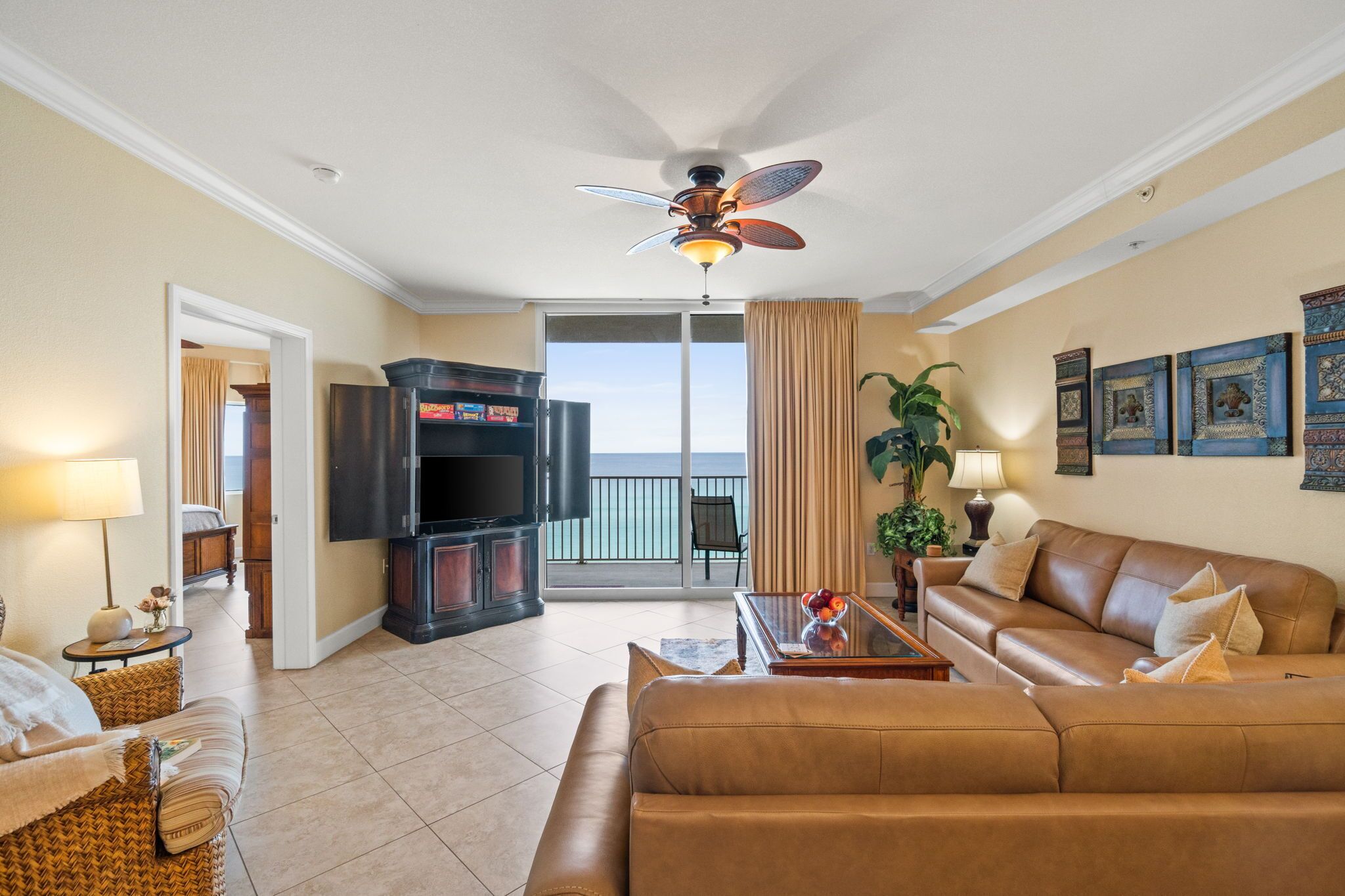 A living room with a sofa, a television, a ceiling fan, and a view of the ocean through a sliding door.