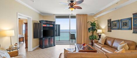 A living room with a sofa, a television, a ceiling fan, and a view of the ocean through a sliding door.