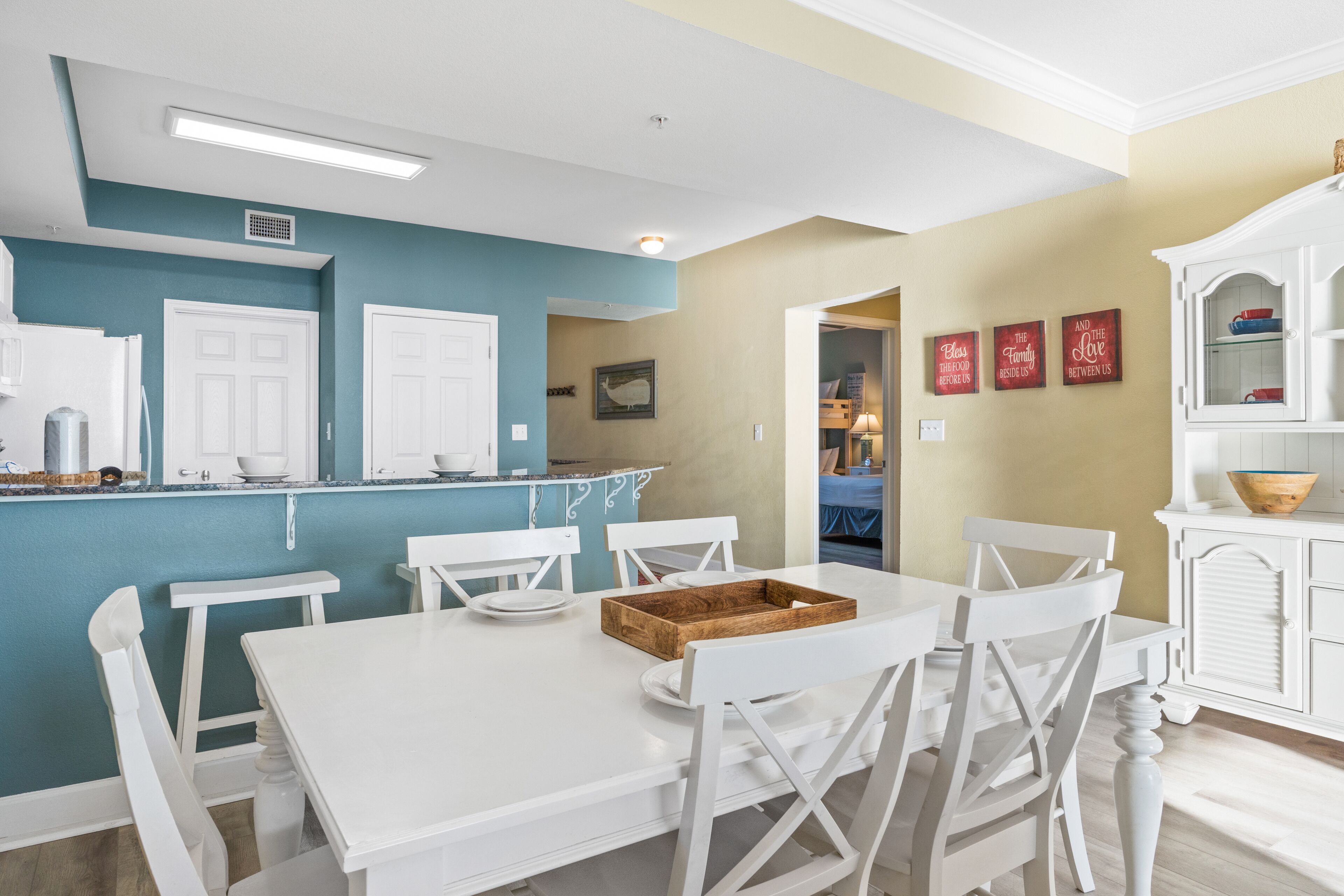 A dining area with a white table and chairs, a wooden tray, and a kitchen in the background.