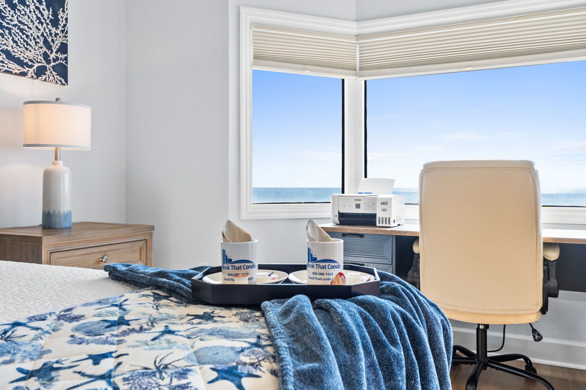 Ein Hotelzimmer mit Bett, Schreibtisch, Stuhl, einem Fenster mit Meerblick und einem Tablett mit zwei Tassen und Taschentüchern.