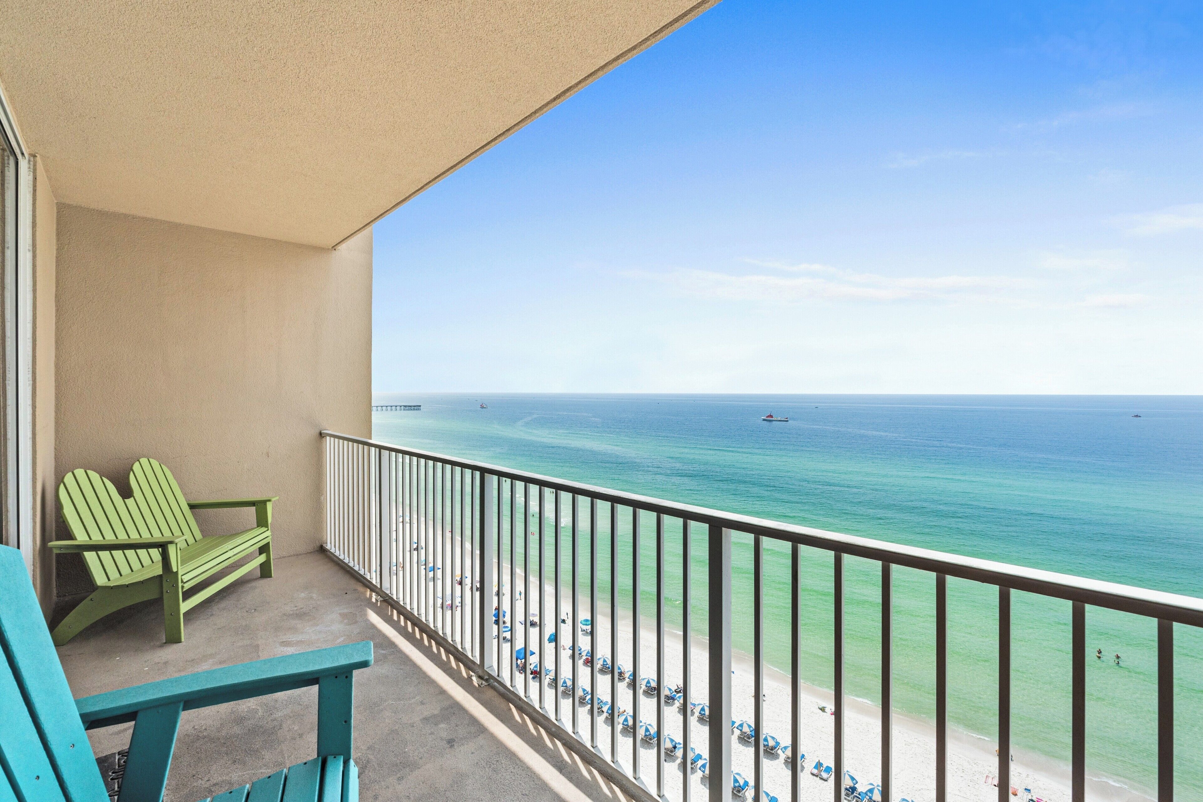 Ein Balkon mit zwei grünen Adirondack-Stühlen, Blick auf den Strand mit Sonnenschirmen und das Meer.