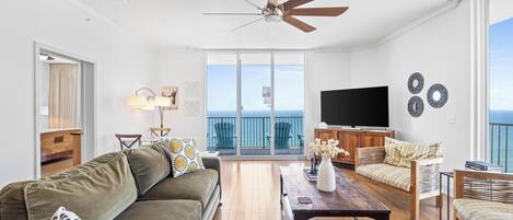 A spacious living room with a ceiling fan, a large flat-screen TV, and a balcony with a view of the ocean.